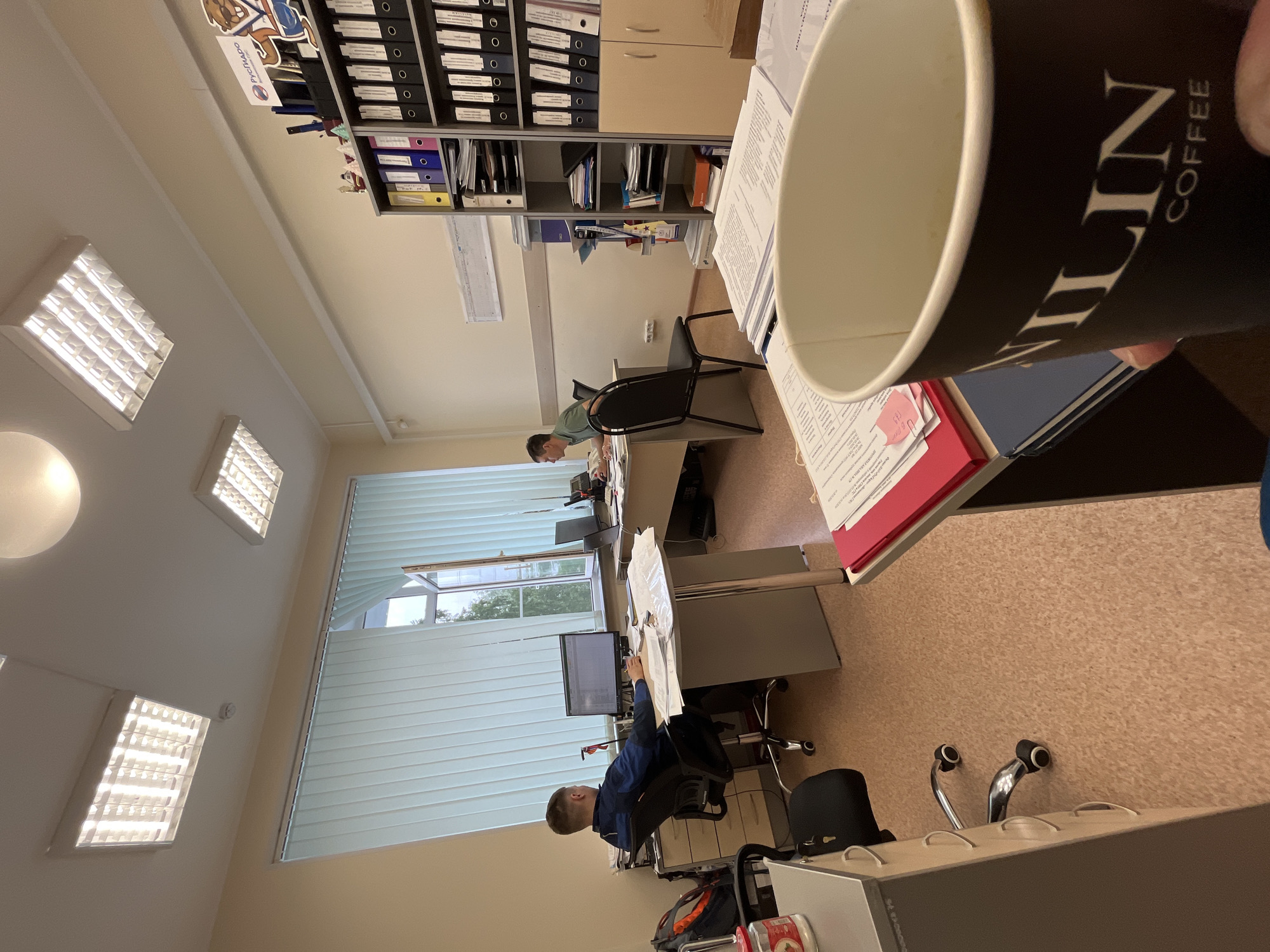 This media file captures an office environment in Chaikovsky, Russia, taken from a slightly elevated, first-person perspective with a black coffee cup held in the immediate foreground. The image is rotated 90 degrees clockwise.
The scene depicts a functional office space with light-colored walls and a light brown floor, possibly linoleum. Fluorescent lighting fixtures illuminate the ceiling, supplemented by natural light from a window with white horizontal blinds. Through the window, green foliage and an overcast sky are visible, suggesting daytime.
Two men are working at separate desks within the office. One man, wearing a green t-shirt, is seated at a desk further back, facing a computer monitor. Closer to the window, another man in a blue jacket is seen from the back and side, also engrossed in work at his computer.
The office is equipped with standard furniture: office chairs, grey desks, and extensive shelving units filled with numerous binders in various colors (black, blue, yellow, red/pink). Papers and documents are scattered on desks, indicating active work. A red binder with a pink sticky note is on a desk in the mid-ground. Near the man in the blue jacket, a black bag or backpack rests on the floor.
Visible text includes "LIN" and "COFFEE" on the black paper cup held by the photographer. On a sticker on a binder on an upper shelf, the Cyrillic text "РусьТяжПром" (RusTiazhProm), likely a company name (Russian Heavy Industry), is discernible. The overall atmosphere suggests a routine workday.