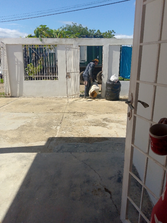 A man, clad in a blue jacket and brown hat, carries a yellow container through an open gate. Behind him are black bags filled with trash, next to a green truck. The scene is framed by a white, weathered wall and a glimpse of the blue sky. The photo is taken from inside a building, with a partially opened door showing a person holding a red cup. The concrete ground is cracked and stained, suggesting wear and age.
