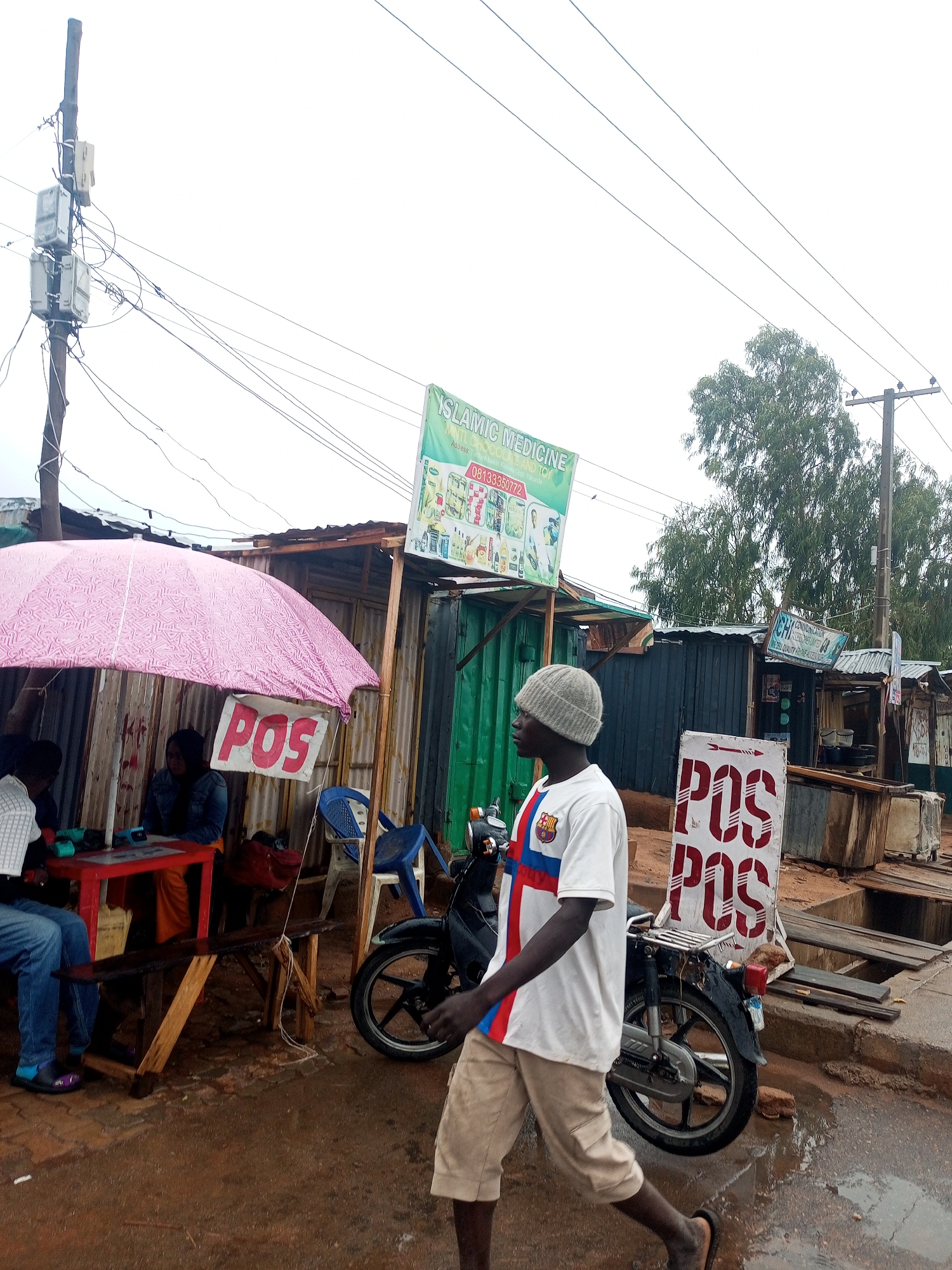 An outdoor scene captures a busy, informal commercial area under an overcast sky. In the foreground, a young man with dark skin, wearing a grey beanie, a white t-shirt with a blue and red cross design, and light-colored shorts, walks past the camera from left to right, his gaze directed forward. He wears flip-flops.
To the left, a vibrant pink umbrella provides shade over a small red table where two individuals are seated. One person, wearing blue jeans, is partially visible on the left, while another, wearing a denim jacket, sits across the table. Behind them are makeshift stalls constructed from corrugated metal sheets and wood. A small sign hangs from the umbrella, prominently displaying "POS" in red letters on a white background.
Further back, to the right of the walking man, a white sign with "POS" repeated multiple times in red letters is visible next to a motorcycle. Beyond this, more rudimentary stalls and structures line what appears to be a muddy or wet dirt road. The ground throughout the visible area is damp and brown.
In the upper-middle background, a green signboard reads "ISLAMIC MEDICINE" at the top, followed by "Adress, Tunlure bak," "ATS AND TOY," "Oppt Yahunde," and a phone number "08133350772." Another smaller sign to the right has partially visible text including "COMMUNICATION" and "NO. SEA QUALITY REPAIRS." Overhead, a wooden utility pole with electrical meters and numerous wires stretches into the bright, featureless sky. Another utility pole and trees are visible further in the distance on the right.