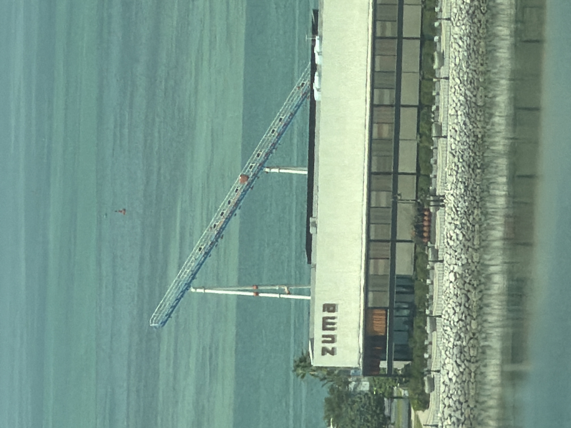 A wide-angle, high-angle shot captures a building with the word "ZUMA" on its facade, situated by the water. A large, inclined structure, possibly a ramp or a conveyor belt, extends from a white support tower towards the right side of the frame, over the water. The building is modern, featuring a row of large windows and a stone-lined waterfront. The water is a vibrant turquoise color, with a distant buoy visible in the background. Lush green trees are visible to the left of the building. The lighting suggests it is daytime.