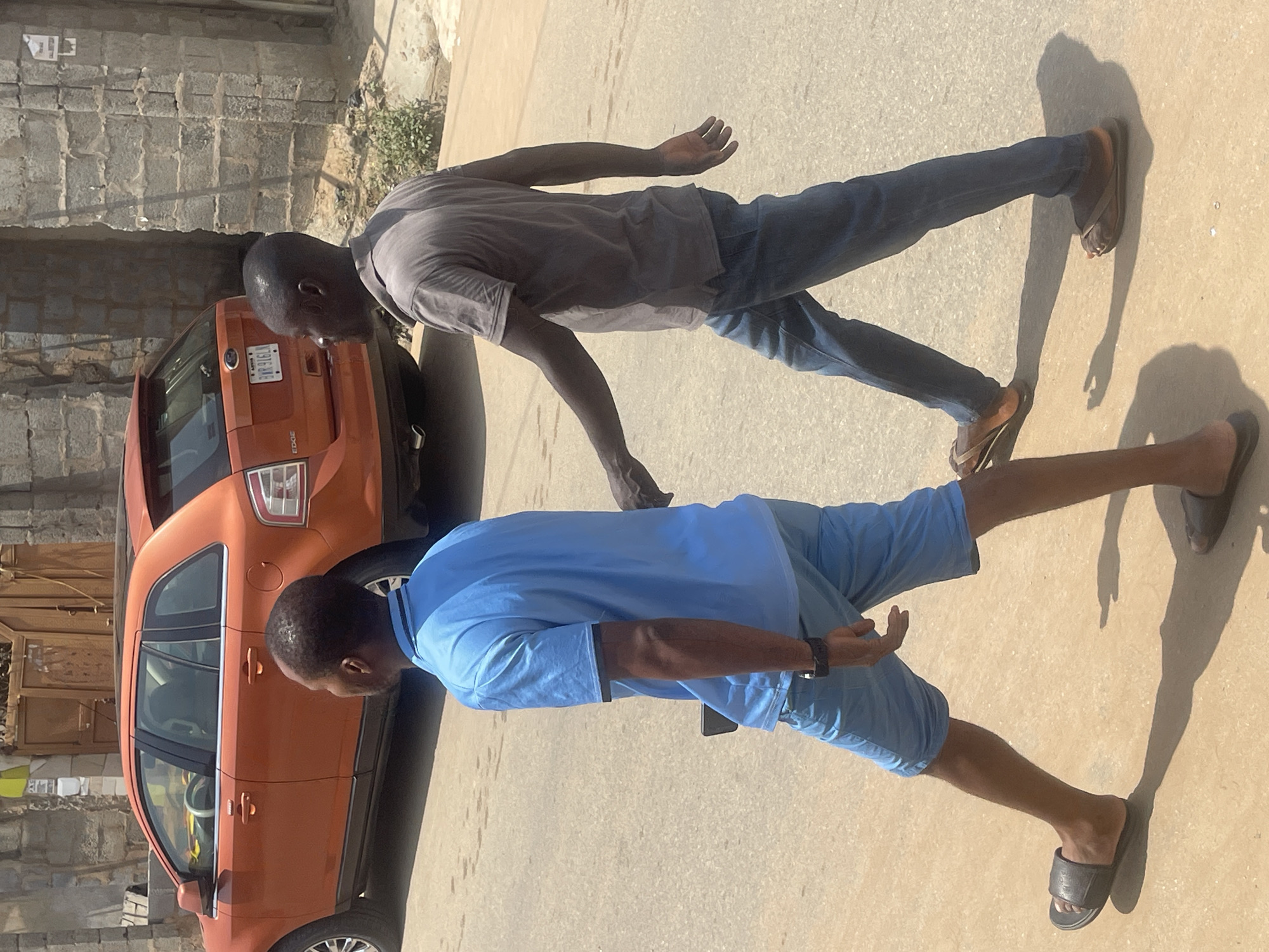 Two men are walking on a paved street in Abuja, Nigeria. The man on the left is wearing a light blue polo shirt and light blue shorts. He has on black slide sandals and is looking to his left. The man on the right is wearing a grey short-sleeved t-shirt and blue jeans. He has on brown flip-flops and is looking to his left. Behind the men, an orange Ford Edge SUV is parked on the street. The background includes a stone wall and a doorway. The sun is casting shadows on the ground.