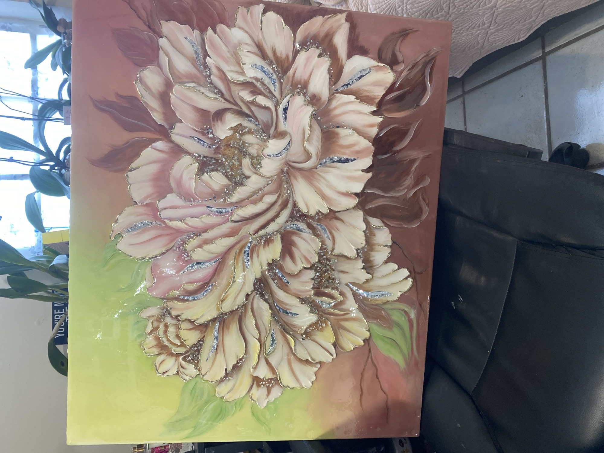 The image shows a painting of a large, multi-petaled flower with a textured, three-dimensional effect. The flower's petals are painted in shades of light pink, cream, and yellow, with accents of gold and silver glitter or small reflective elements. The background of the painting transitions from a bright yellow-green on the left to a dusty rose or brown on the right, with darker brown and reddish-brown leaf-like shapes. The painting appears to be displayed indoors, possibly on a wall or easel, as it is positioned above a black leather chair and a tiled floor. In the background, a window with houseplants is visible. There is no specific indication of the city of Miami, United States.