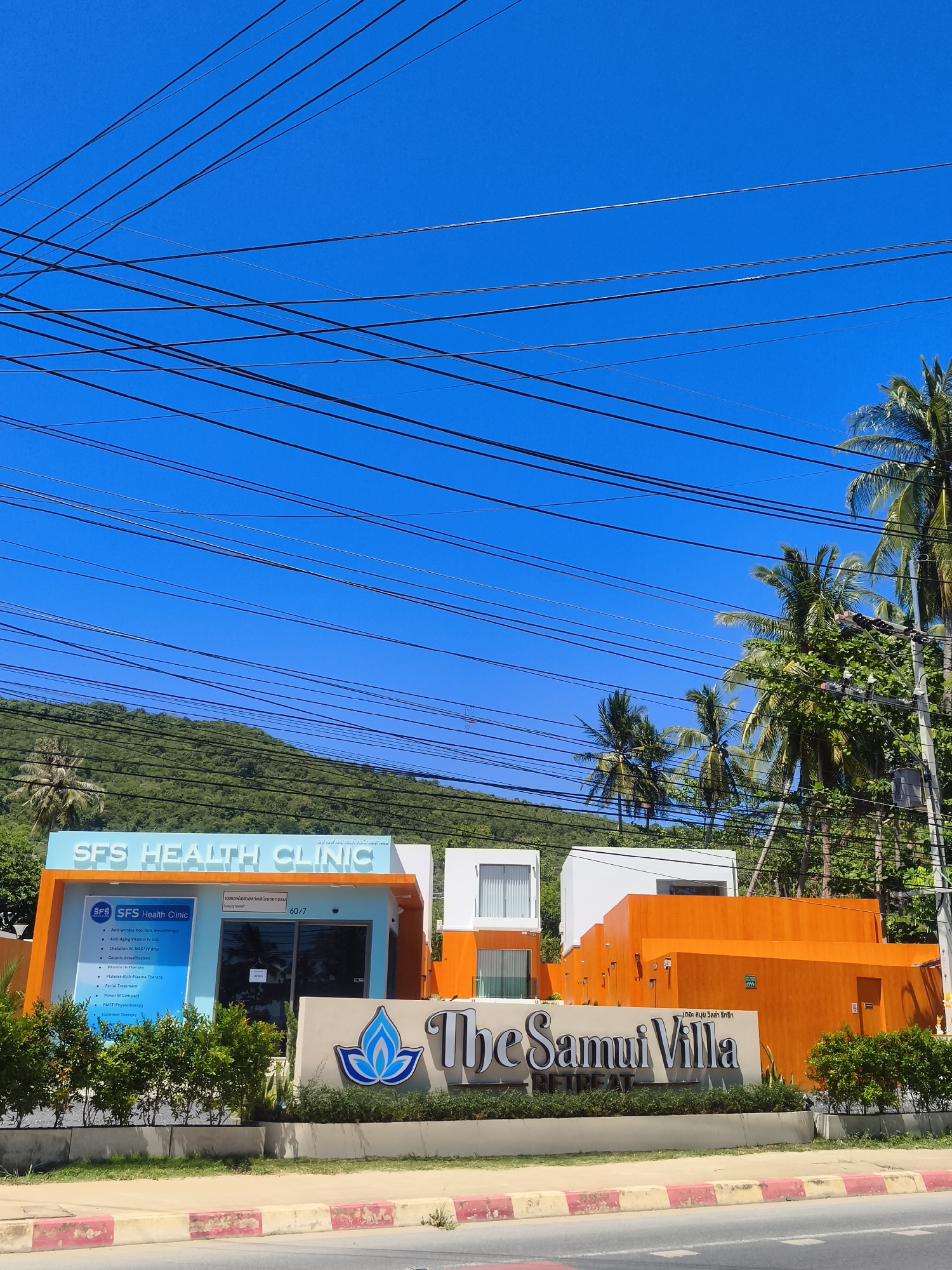 The image shows a sunny exterior view of a modern building complex in Baan Thong Po, Thailand. The main focal point is "SFS Health Clinic," a bright blue and orange building with a sign listing various services. To the right of the clinic, a larger, vibrant orange structure is visible, which appears to be part of "The Samui Villa Retreat," indicated by a large sign. Palm trees and lush green foliage surround the buildings, with a verdant hillside in the background. Overhead, a complex network of power lines crisscrosses the clear blue sky. No people are visible in the scene. The street in front of the complex is paved with red and white curbs.