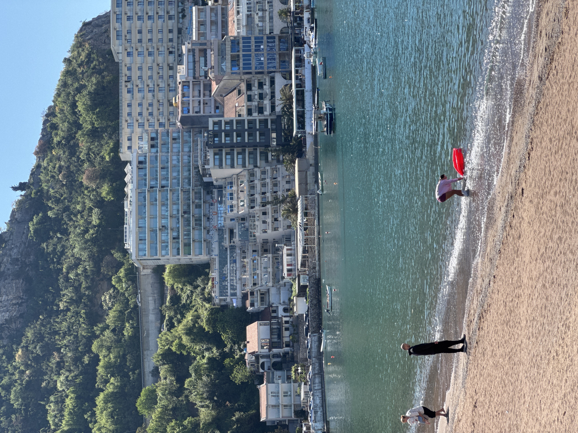 The image depicts a view of Rafailovići, Montenegro. The scene includes a sandy beach bordering turquoise water. On the beach, three people are visible: One person is standing near the water's edge. A second person is farther away and appears to be walking along the shoreline. A third individual is at the water with a red flotation device. In the background, there are multiple buildings, some of which are high-rise apartments, nestled against a tree-covered mountain. Several boats are visible docked along the waterfront.