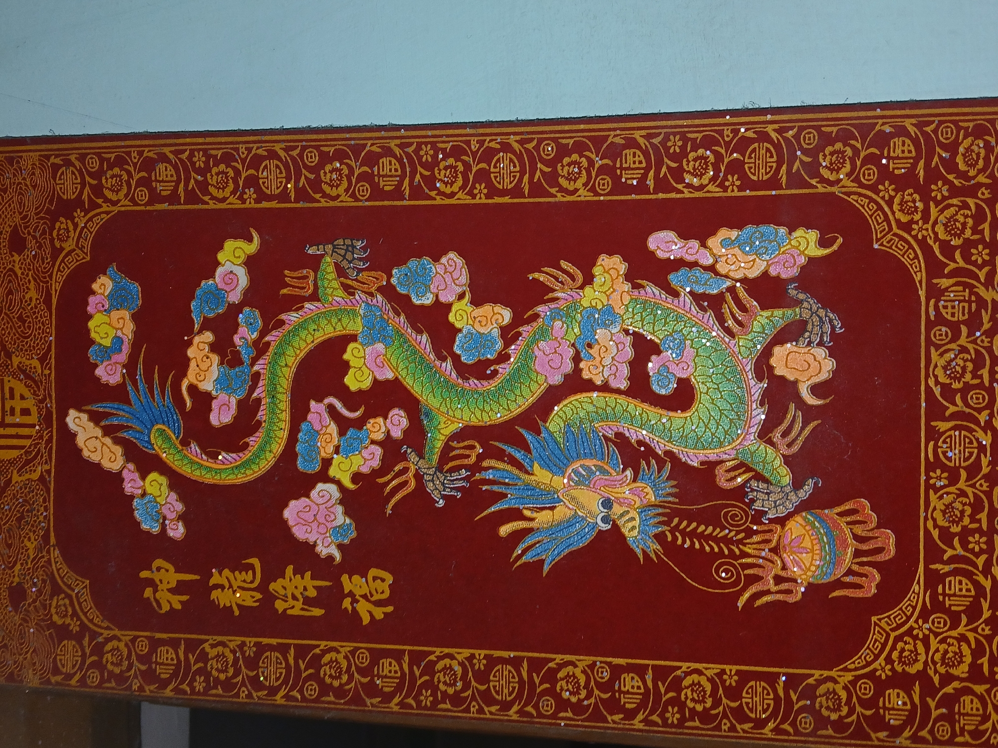 A photograph captures a rectangular, maroon-colored surface adorned with a golden border featuring floral and geometric patterns. Centered on this surface is an intricately designed green dragon, surrounded by colorful clouds. Below the dragon are yellow Chinese characters. The image was taken in Bengaluru, India.
