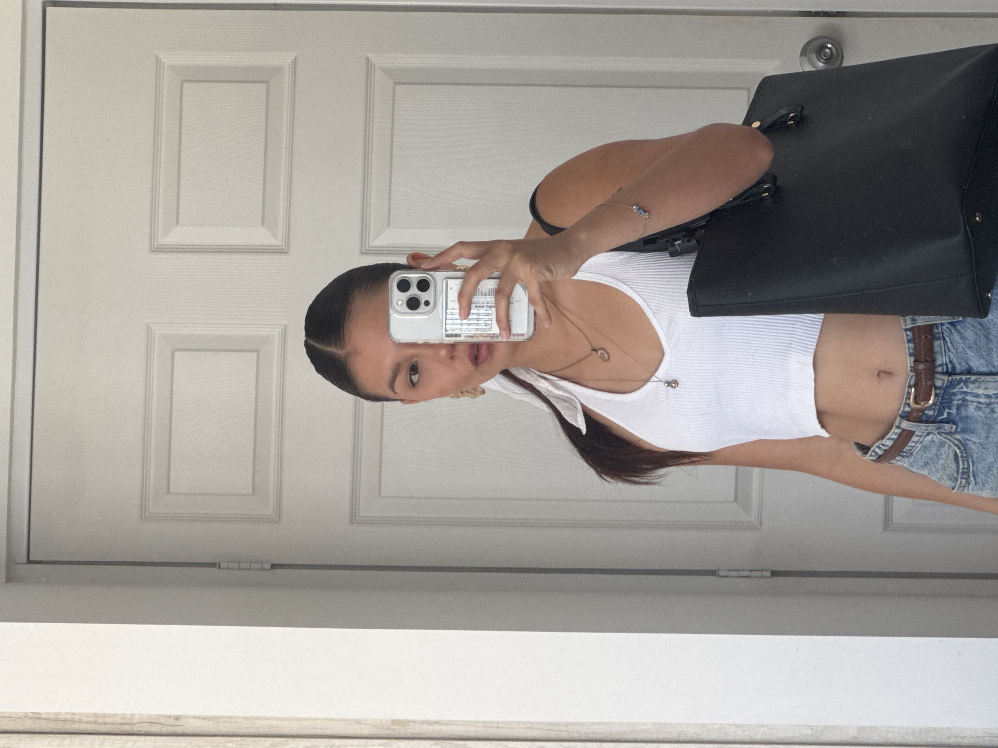 A young woman is taking a selfie in front of a white paneled door. She's wearing a white tank top and blue denim shorts with a brown belt. She's holding a smartphone with a clear case in front of her face. A black tote bag hangs from her shoulder. The location appears to be indoors. The city of San Francisco Ocotlán (Ocotlán), Mexico is not discernible from the image.