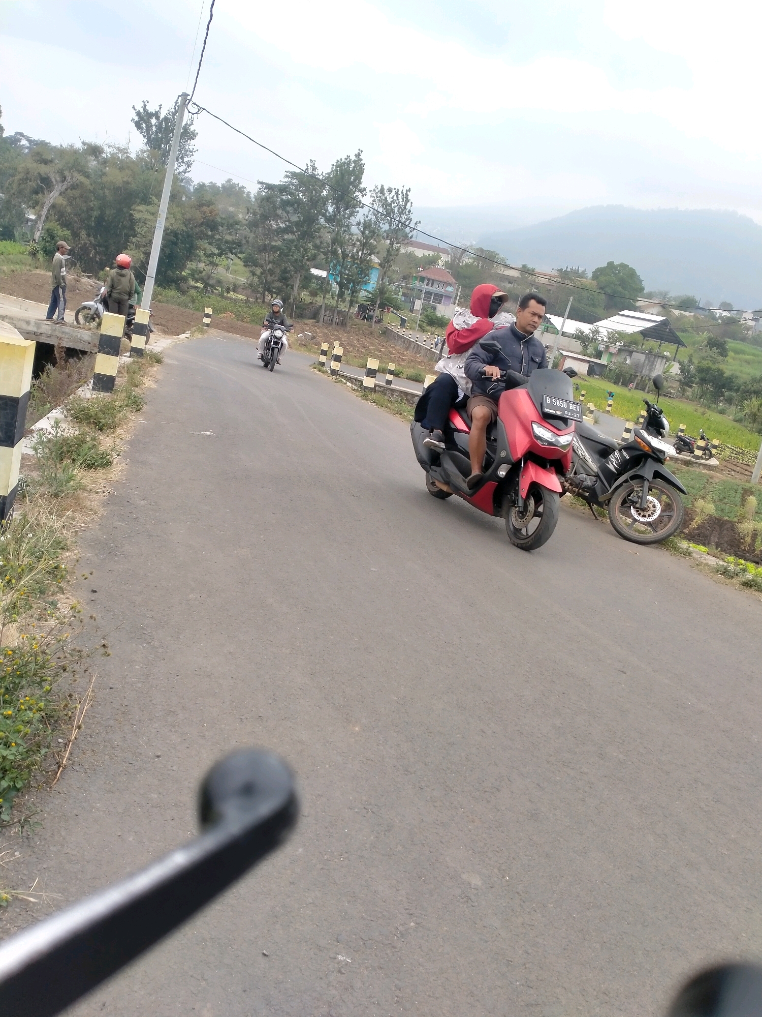 In the city of Batu, Indonesia, on a paved road that winds through a hilly, green landscape, a red motorcycle carrying two individuals is prominently featured. The male driver, wearing a blue jacket and brown shorts, is looking forward. Seated behind him is a female passenger wearing a red hooded jacket and dark pants. They are accompanied by a black motorcycle parked to their right. In the background, a few houses and scattered trees dot the terrain, with a hazy mountain range forming the horizon. Several people are visible on the side of the road, some with motorcycles, suggesting a possible stop or gathering. The scene is illuminated by natural daylight under a cloudy sky.
