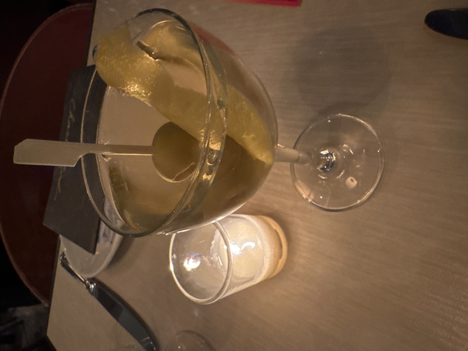 An overhead, close-up view depicts a dimly lit indoor setting, likely a restaurant or bar within New York, United States of America. The primary subject is a clear martini glass containing a translucent, pale yellow beverage, a large pickle spear, and a green olive speared by a white, flat-headed toothpick. To the left of the martini glass, a small clear glass votive holder contains a lit white candle, casting a soft, warm glow on the light-colored table surface. In the background, partially visible, are a silver-bladed knife, a white plate with a dark menu or card resting on it, and a dark red upholstered chair back. No individuals are present in the frame; the scene is static.