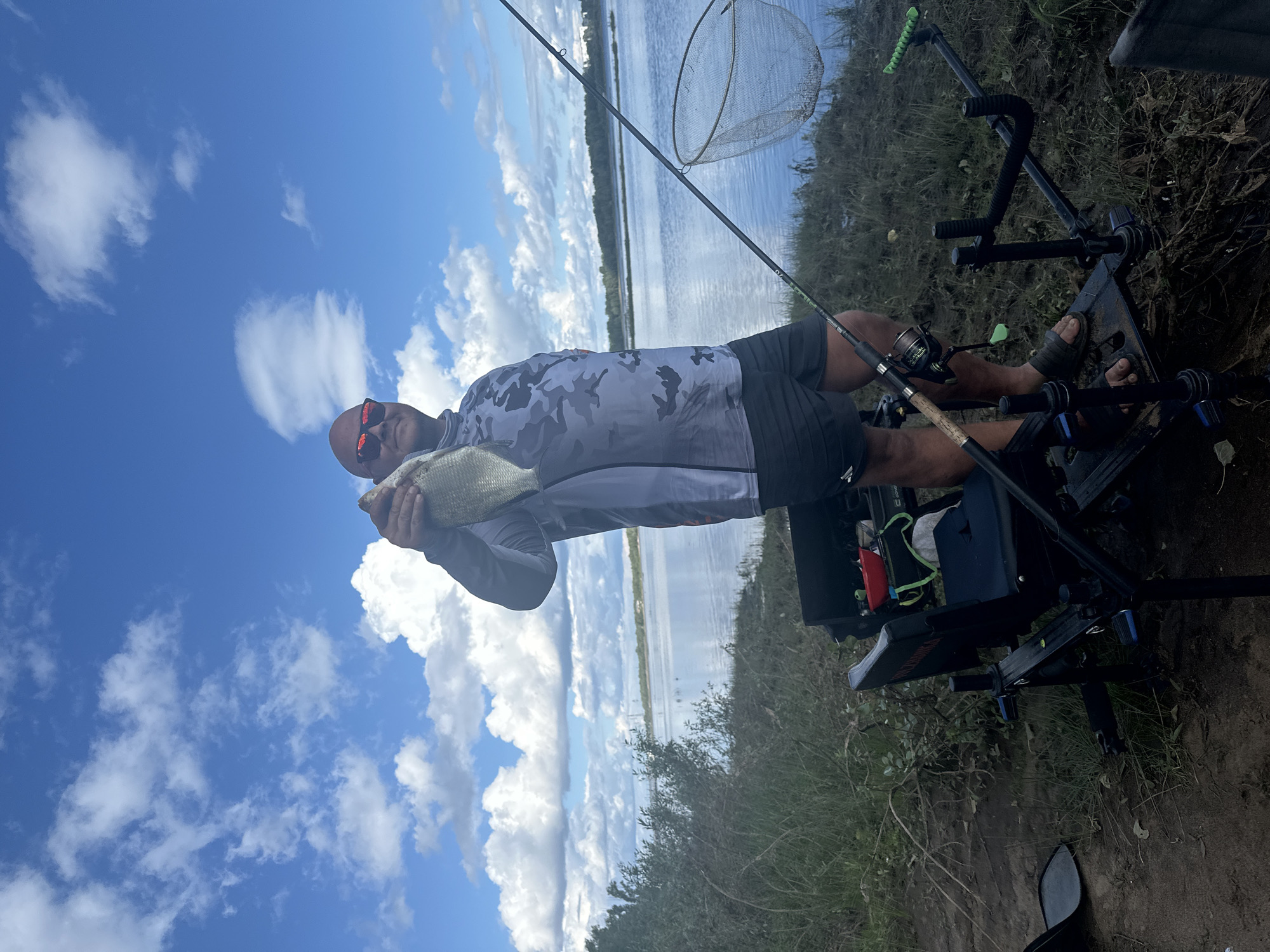 A bald man wearing sunglasses and a gray camouflage long-sleeved shirt and black shorts is holding a freshly caught fish near a body of water, likely a river, on a cloudy day. He is standing next to fishing equipment, including a rod holder and a fishing net. The scene appears to be a rural or natural setting, possibly on the outskirts of a city. The man is smiling, suggesting satisfaction with his catch. The background shows a calm body of water with trees lining the opposite bank and a cloudy sky overhead. The overall impression is one of a successful fishing trip in a serene natural environment.