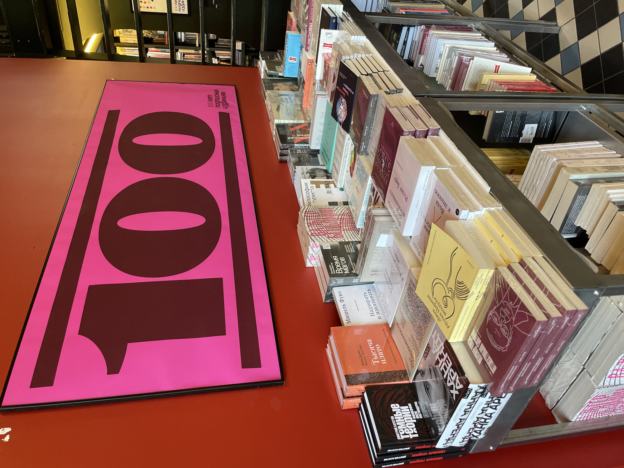 Here is a detailed description of the image:
The image shows a bookstore interior, with a bright red table surface in the foreground. A large pink poster with black text (Russian, saying "100 years of subscription editions") and bold numerals "100" printed on it is placed on the table.
Next to the poster, a metal shelf unit is filled with books. The books are arranged in rows, standing upright and closely packed together. Many book covers are visible, displaying titles and author names in Russian. There is a variety of colors and designs on the book covers.
In the background, other shelves filled with books can be seen. The flooring is tiled with black and white squares, and the lighting appears to be a combination of natural and artificial. The overall environment suggests a well-stocked bookstore.
Based on the Cyrillic text and general aesthetics, the location is likely Saint Petersburg, Russia, as indicated by the "100 years of subscription editions" text often associated with publishing houses in the city.