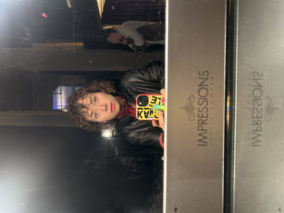 A young person with curly brown hair and a red bandana around their neck takes a selfie in a mirror. They wear a black leather jacket and hold a phone with a brightly colored case. In the background, a dimly lit room with a window is reflected. Another person lies on a surface in the upper portion of the mirror's reflection. The side of the mirror has the words "IMPRESSIONS VANITY COMPANY" printed vertically in gray lettering.
