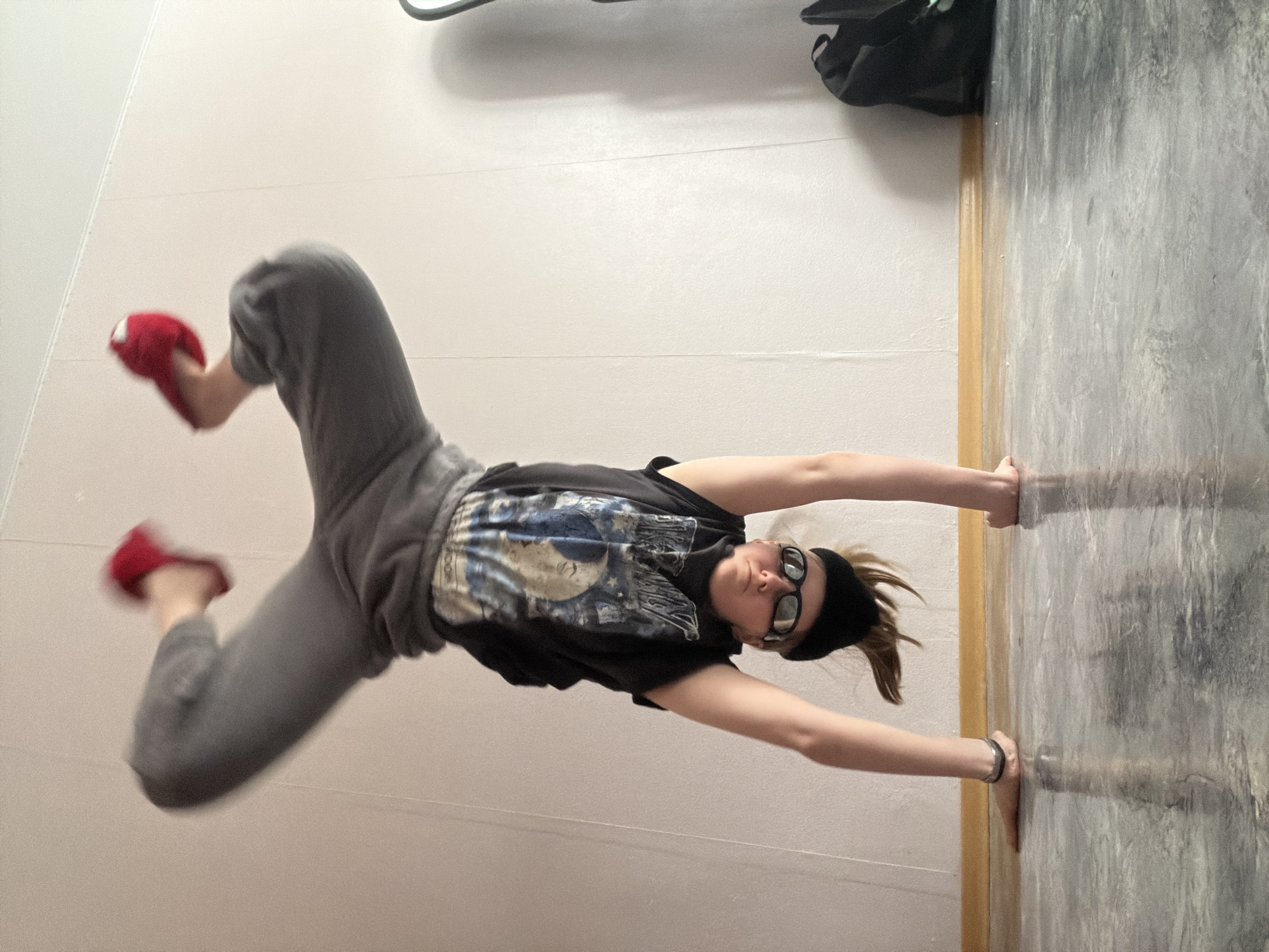 A person wearing gray sweatpants, a black T-shirt, glasses, a black headband, and red slippers is doing a handstand with their hands against a wall. The background consists of a white wall and a gray wall with a wood trim. There is a black bag visible near the top of the frame. It is impossible to determine the location within the city of Perm, Russia from this image.