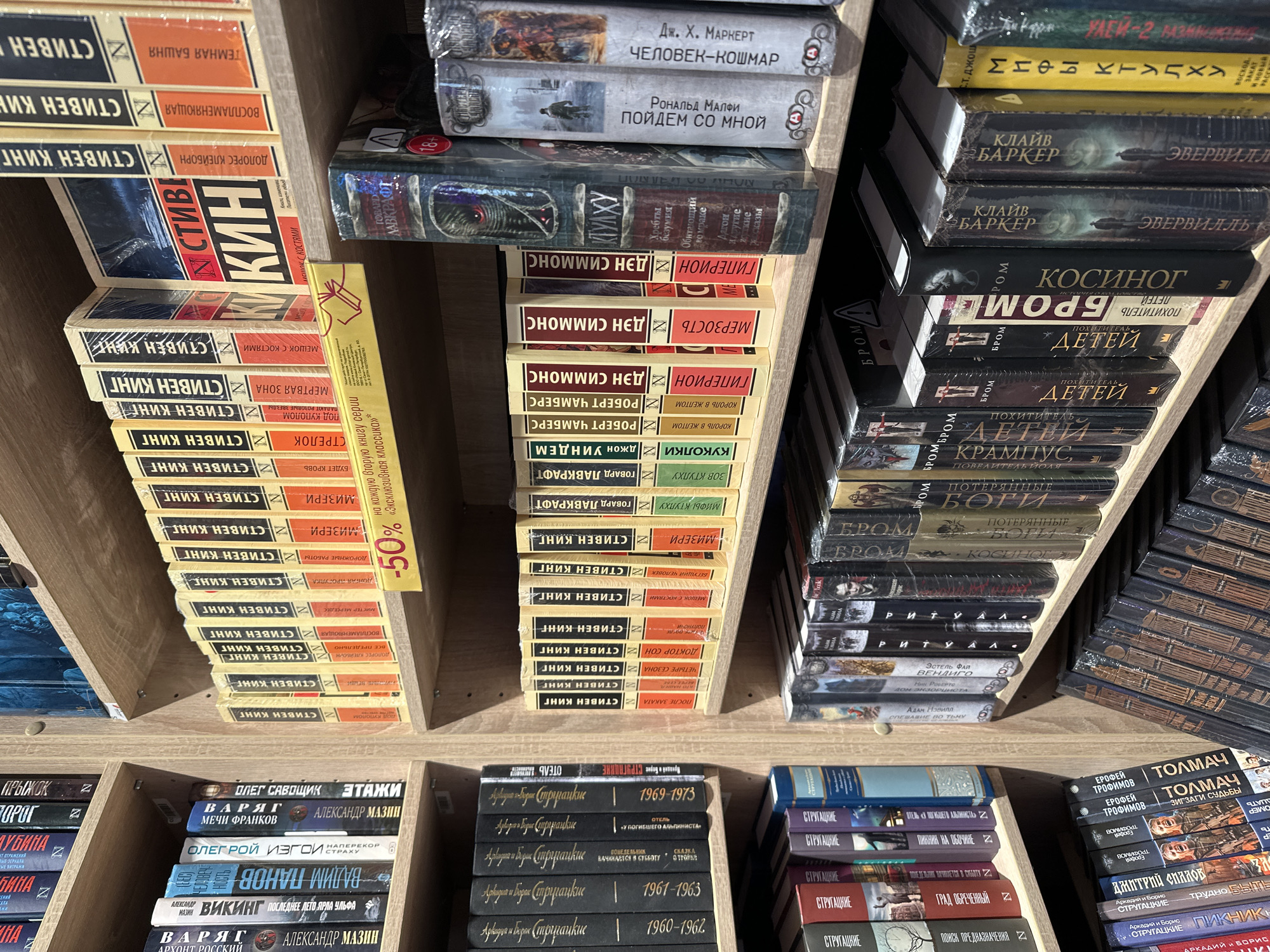 The image shows a close-up view inside a bookstore, likely in Kazan, Russia, given the Cyrillic text on the book covers. The shelves are filled with neatly stacked books, mainly paperback editions.
The books cover various genres, including horror, science fiction, and fantasy, with several titles by Stephen King, Clive Barker, Dan Simmons, and others. There's a yellow price tag indicating a 50% discount on the second book in a series. The books are arranged on light-colored wooden shelves, viewed from above. The lighting is bright, but some books appear shadowed.
The books are placed in a store and available for sale.