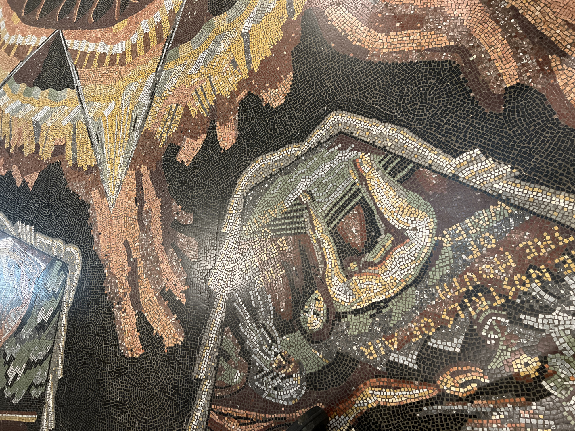 The media file showcases a detailed section of a large mosaic artwork, composed of numerous small, irregularly shaped tesserae. The predominant color palette consists of earthy tones including various shades of brown, cream, yellow, gray, and muted green, set against a black background.
The mosaic depicts a dynamic and stylized scene. In the upper and central parts, large, abstract or organic forms resembling wings or feathered patterns are visible, characterized by sweeping lines and triangular sections in warm browns, yellows, and creams. Towards the center-right, a human-like figure is rendered in a contorted or active pose, outlined in lighter tesserae of cream, gray, and muted green. A distinctly stylized hand with fingers is visible, appearing to reach or grasp. Another suggested limb or form is present at the bottom right.
The overall composition is highly stylized, employing flowing lines and geometric elements within the larger forms. The stark black tesserae of the background provide strong contrast, causing the figures and patterns to prominently stand out. The artwork appears to be a large-scale decorative piece, possibly a floor or wall mosaic, exhibiting an Art Deco or similar early 20th-century aesthetic. No text is visible in the provided images. The location of the artwork is London, United Kingdom.