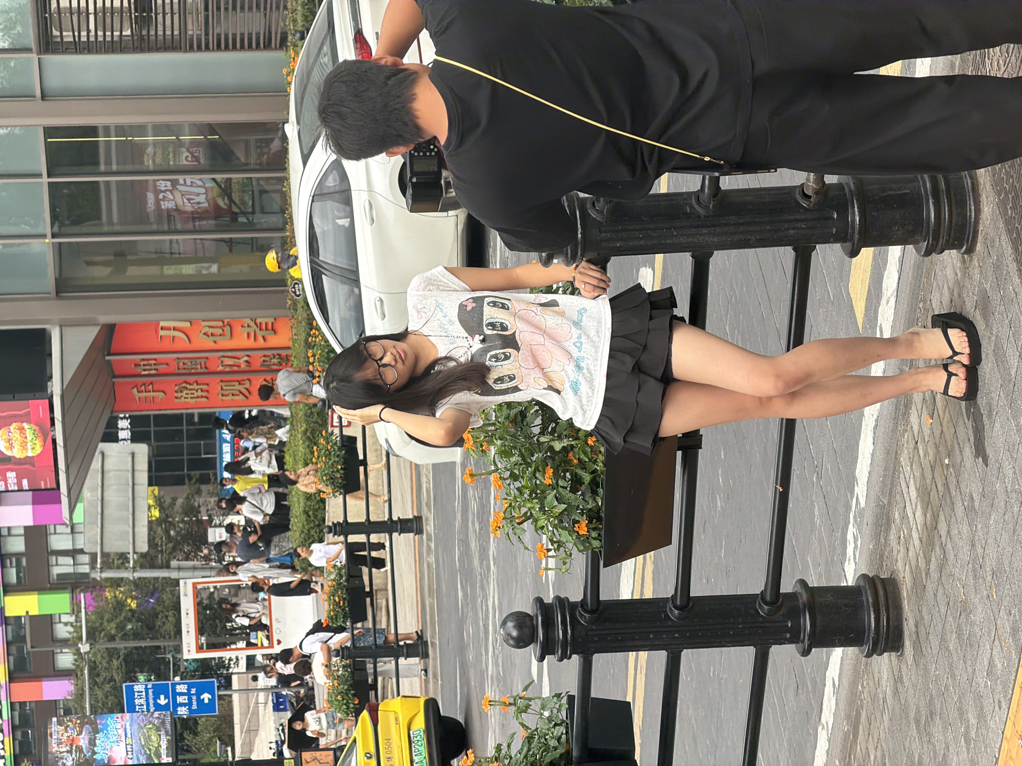 In Yuzhong District, China, a young woman with glasses wearing a white t-shirt with a cartoon print and a black ruffled skirt stands by a black metal fence. She is holding her hand to her head, and her legs are visible below the skirt, with black flip-flops on her feet. To her right, a person with short dark hair, wearing a black t-shirt and pants, is facing away from the camera, holding a camera and appearing to take a photo of the woman. In the background, a white car is parked behind the fence. Beyond the car, buildings with colorful facades are visible, along with other pedestrians and signage. A yellow car is parked on the left side of the frame. The scene takes place on a street with pavement and a sidewalk, and planters with orange flowers are situated along the fence.