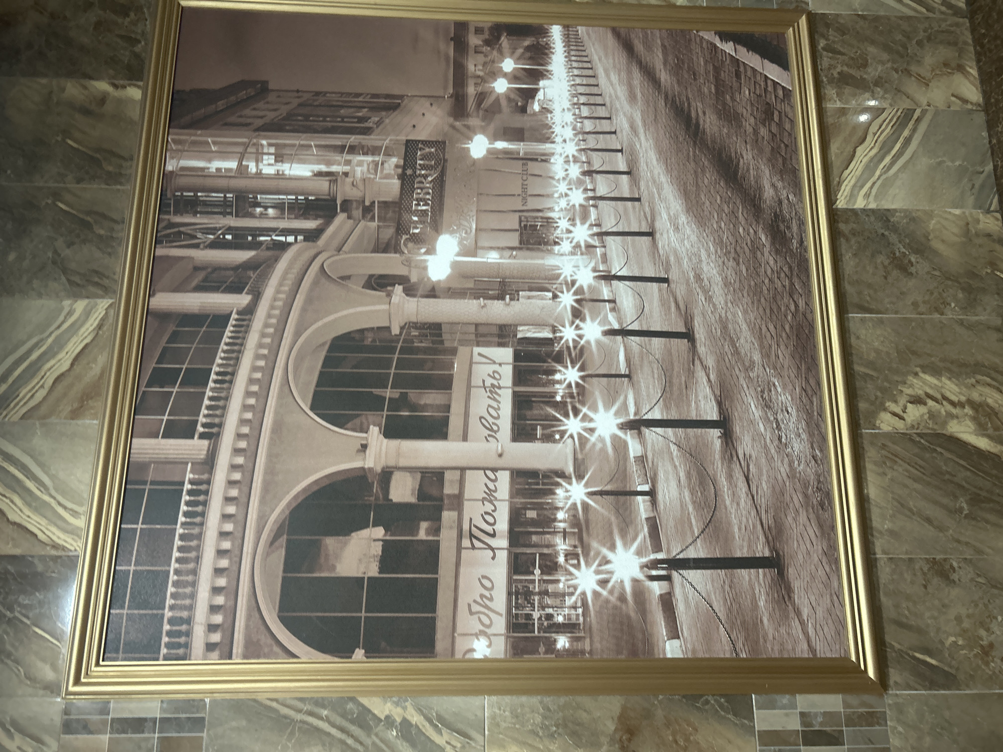 The image shows a framed sepia-toned photograph of a street scene at night, likely a commercial area, possibly a club or restaurant. The photograph is hanging on a wall covered with marbled tiles. The scene depicts an arched building facade with large windows and a prominent sign that reads "CELEBRITY NIGHT CLUB". A row of lampposts with starburst lights lines a cobblestone street, and bollards with chains create a barrier along the sidewalk. There is text in Russian on the building facade that appears to read "Добро Пожаловать", meaning "Welcome". The overall impression is one of a lively night entertainment district.