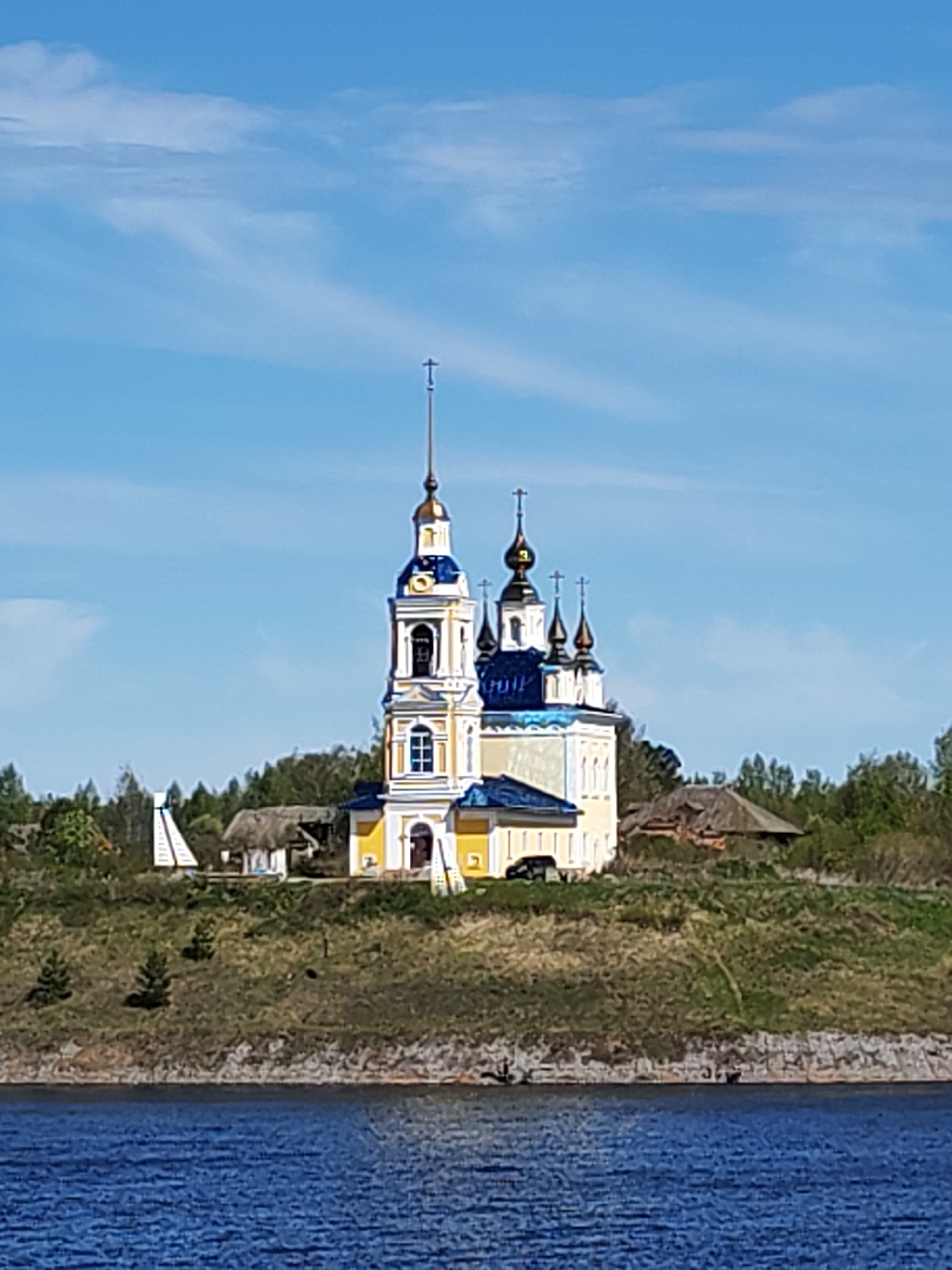 The image shows a yellow and white Russian Orthodox church with blue domes, located in Глебово, Russia. The church sits atop a grassy hill overlooking a body of water. There are trees and other buildings visible around the church, including a small, white, tower-like structure to the left of the church. A black car is parked near the church. The sky is blue with some scattered white clouds.