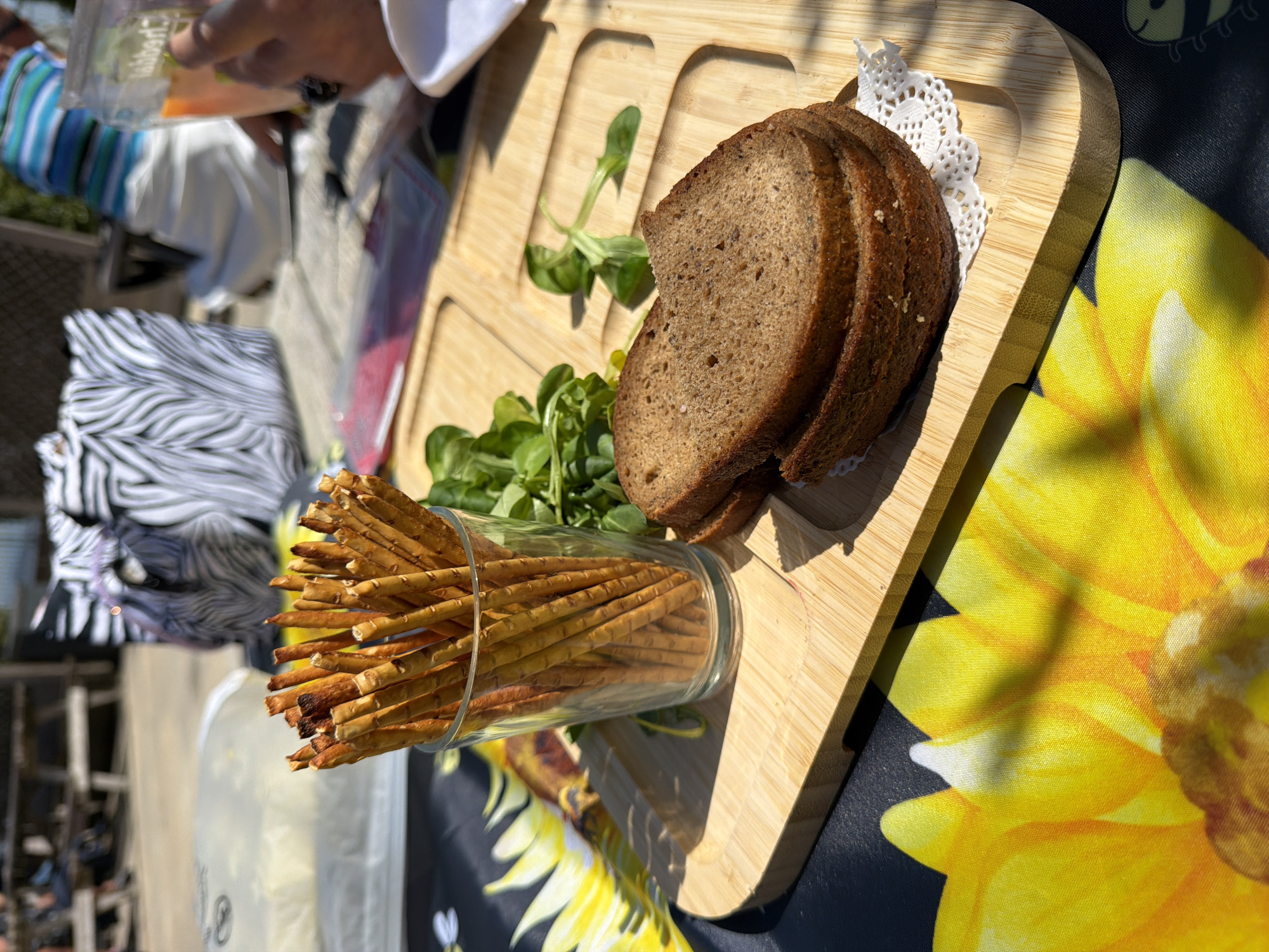 The image displays an outdoor setting in bright daylight, likely at a casual gathering in Monistrol de Montserrat, Spain. The main subjects are food items arranged on a table.
In the foreground, a light-colored wooden serving board with carved compartments holds several slices of dark bread, placed on a white doily. Beside the bread, sprigs of green leafy garnish, possibly lamb's lettuce, are spread. To the left of the board, a clear glass is filled with numerous thin pretzel sticks or breadsticks.
The serving board rests on a black tablecloth featuring a large, vibrant yellow sunflower design. Strong shadows cast by the board and the food indicate bright, direct sunlight, suggesting the time of day is midday or early afternoon.
In the upper left background, a person's hand holds a clear glass or cup. Faint text, appearing to read "Vibert" or "Hibert," is visible on this glass. Further in the background, a chair with a distinctive black and white zebra-stripe pattern is partially visible. The background elements are blurred, suggesting an outdoor environment with other structures. The scene depicts a relaxed meal or snack being enjoyed outdoors.