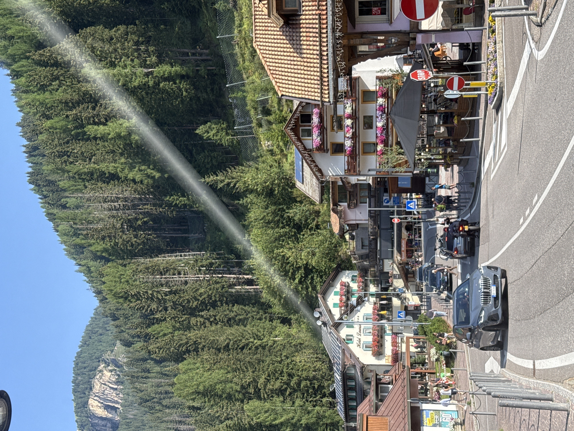 The image shows a street scene in Canazei, Italy, a town nestled in the Dolomites mountain range. In the foreground, a gray BMW SUV is driving down a paved road. To the right of the SUV, a black motorcycle with a rider is visible. The road is lined with buildings, some of which appear to be hotels or restaurants, decorated with flowers on their balconies. A black umbrella shades an outdoor seating area. Traffic signs, including a stop sign, are present. In the background, a steep, forested mountainside rises, with a rocky outcrop visible near the top. Sunlight streams through the trees, creating a visible beam of light. People are walking on the sidewalks and in the street.