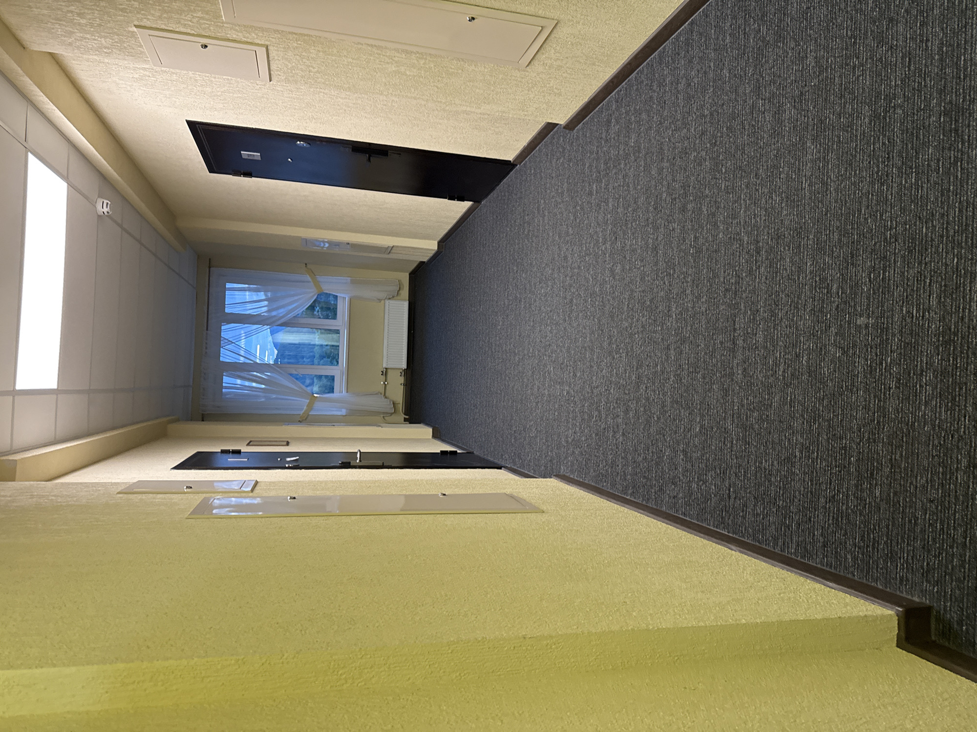 The image shows a long, carpeted hallway with yellow textured walls. The hallway is dimly lit by overhead fluorescent lights and natural light coming from a large window at the far end. The window has sheer white curtains and offers a view of a slightly overcast sky and some greenery. There are several dark doors visible along the hallway, one on the right and others on the left which are partially obscured. Wall-mounted electrical panels or utility boxes are present on the walls. A white radiator is visible beneath the window. The floor is covered with a dark grey, patterned carpet. The scene appears to be a corridor in a residential or commercial building in Syrostan, Russia. There are no people or animals visible in the image.