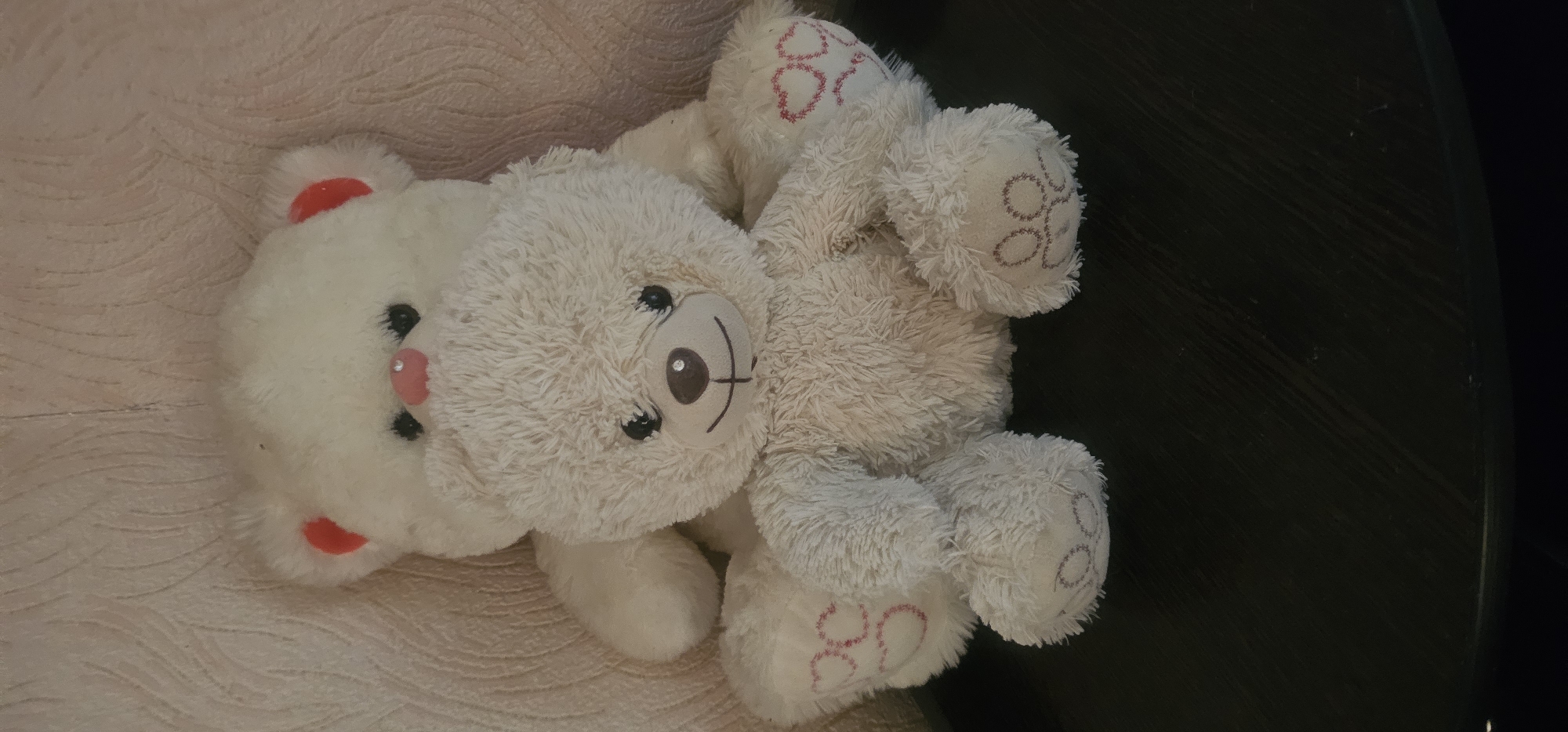 Two white teddy bears are positioned in the foreground and background, seated on a dark, textured surface. The front teddy bear is closer and larger, with a fluffy, off-white fur coat and a brown nose with a light brown embroidered smile and a small, circular, gem-like decoration in the center of its snout. Its eyes are black, round, and shiny. It has red heart shapes embroidered on its paws. The second teddy bear, behind the first, is larger and less detailed, with a more uniform white appearance and pink inner ears. Its nose is pink and its eyes are black. The background is a light-colored, textured wall, possibly wallpaper. The image is taken from a medium-close shot, focusing on the two stuffed toys. The location in Oktiabrskaya, Russia is not discernible from the image.