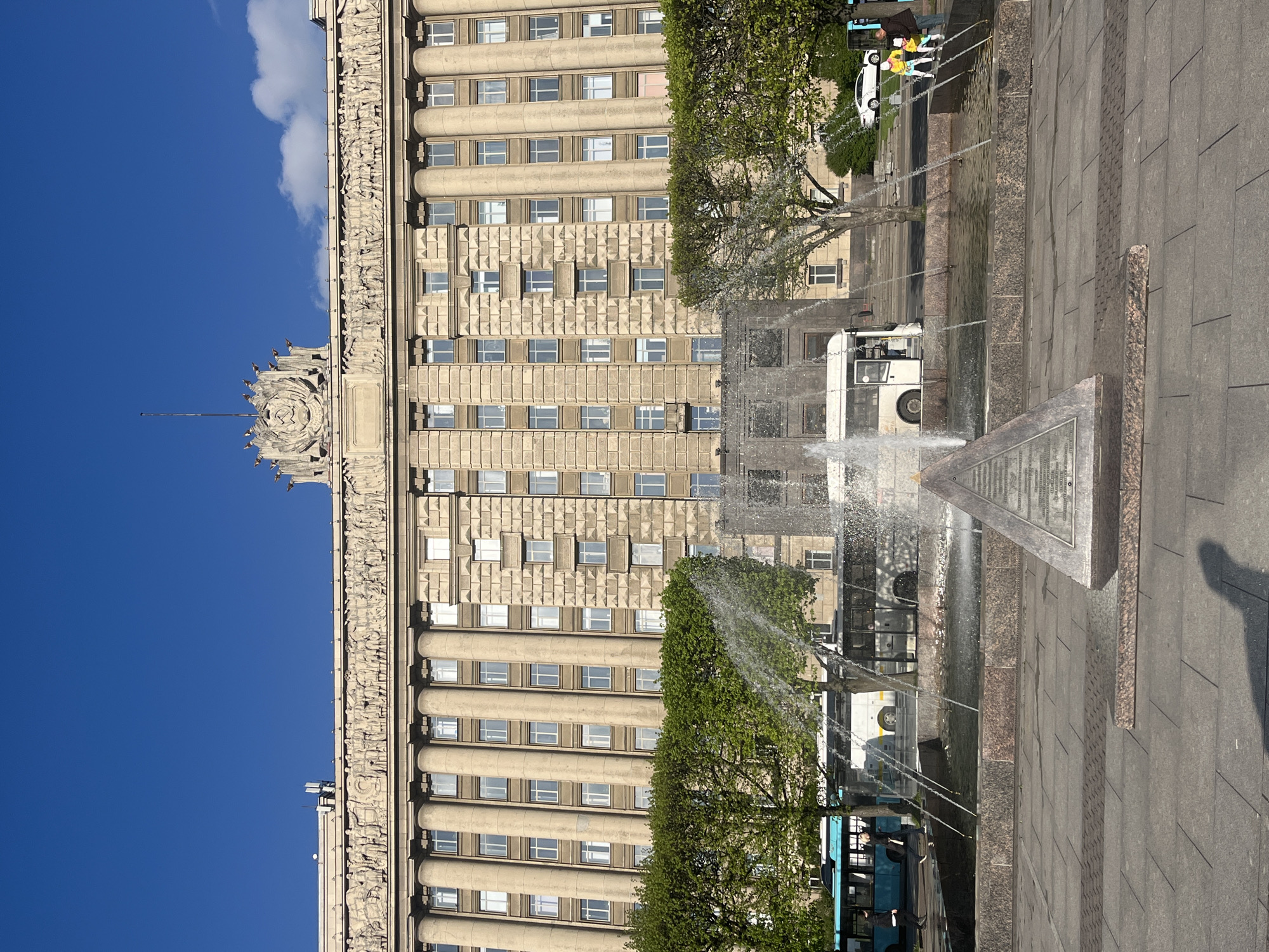 This photo shows the cityscape of Saint Petersburg, Russia. A tall, light-tan-colored building with multiple floors and windows stands out against a clear blue sky. The building has decorative architectural elements, including columns and a detailed frieze at the top. A fountain is positioned in front of the building, with water jets spraying into the air. The fountain seems to have a stone monument or structure at its base. A few buses and cars are visible behind the fountain, along with some trees. People are standing near the vehicles. The ground is paved with large, gray stone slabs. The image appears to have been rotated.