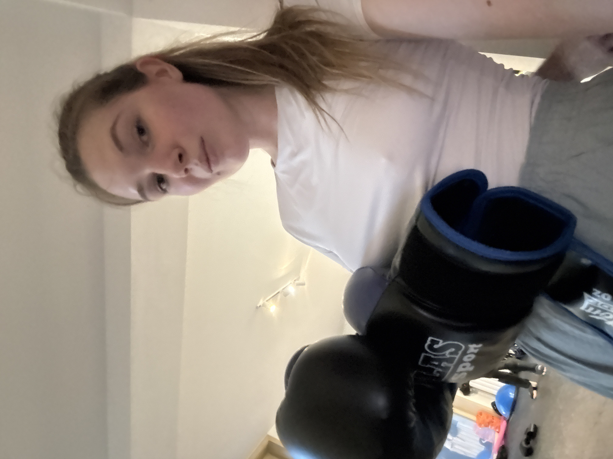 A woman with light skin, brown hair, wearing a white t-shirt and grey sweatpants, is holding up a pair of black boxing gloves with blue trim. The gloves have white lettering and a logo. The background includes a white ceiling with track lighting and a partially visible room with exercise equipment. No specific location in Belgrade, Serbia, can be determined from the image.