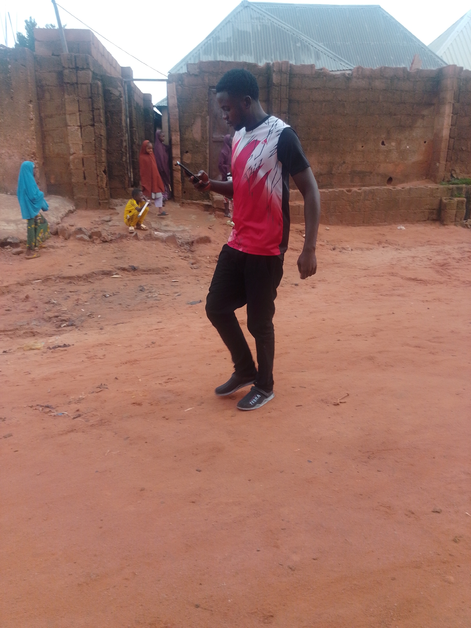In a street scene in Tempure, Nigeria, a young Black man dressed in a red and white patterned t-shirt and black pants stands in the foreground, holding a dark mobile phone. He is looking down at the phone. To the left of the man, a young girl wearing a bright blue headscarf and a patterned skirt stands on a raised dirt area. Further back, two women are visible: one in a rust-colored headscarf and the other in a dark purple headscarf. A child in a yellow outfit is crouched down. The background features buildings constructed with reddish-brown bricks and corrugated metal roofs. The ground is composed of reddish-brown dirt.