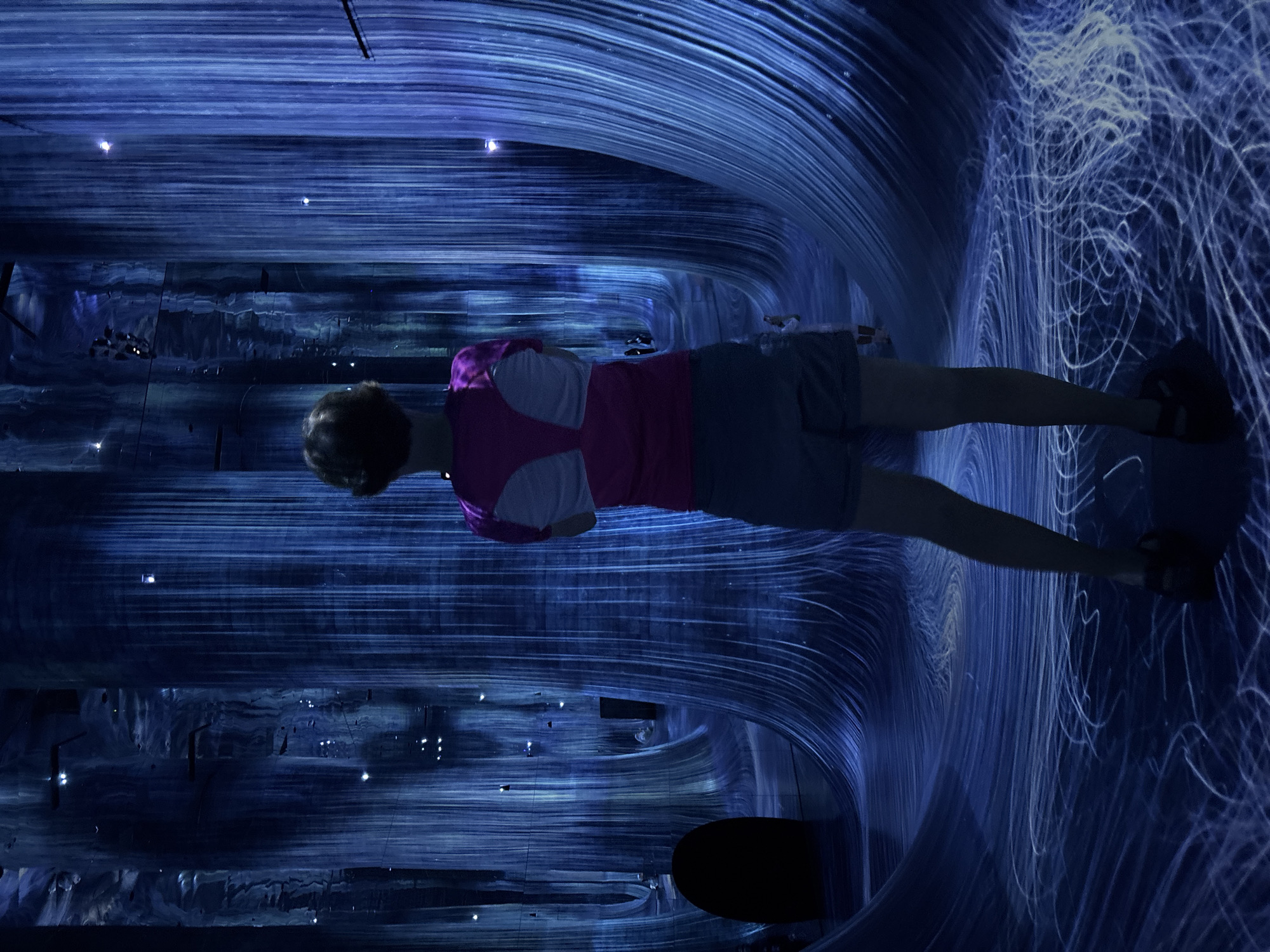 The image captures a person standing in a dimly lit room with a surreal, immersive art installation. The environment is characterized by vertical, illuminated blue lines that create the illusion of a forest or cascading water. The floor also features swirling patterns of light. The person, seen from behind, is wearing a pink and white t-shirt and grey shorts, with their arms crossed. The overall atmosphere is tranquil and otherworldly, suggesting a contemporary art exhibition. While the image is a high-resolution capture, it does not provide any specific geographical markers to identify its location within Abu Dhabi, United Arab Emirates.