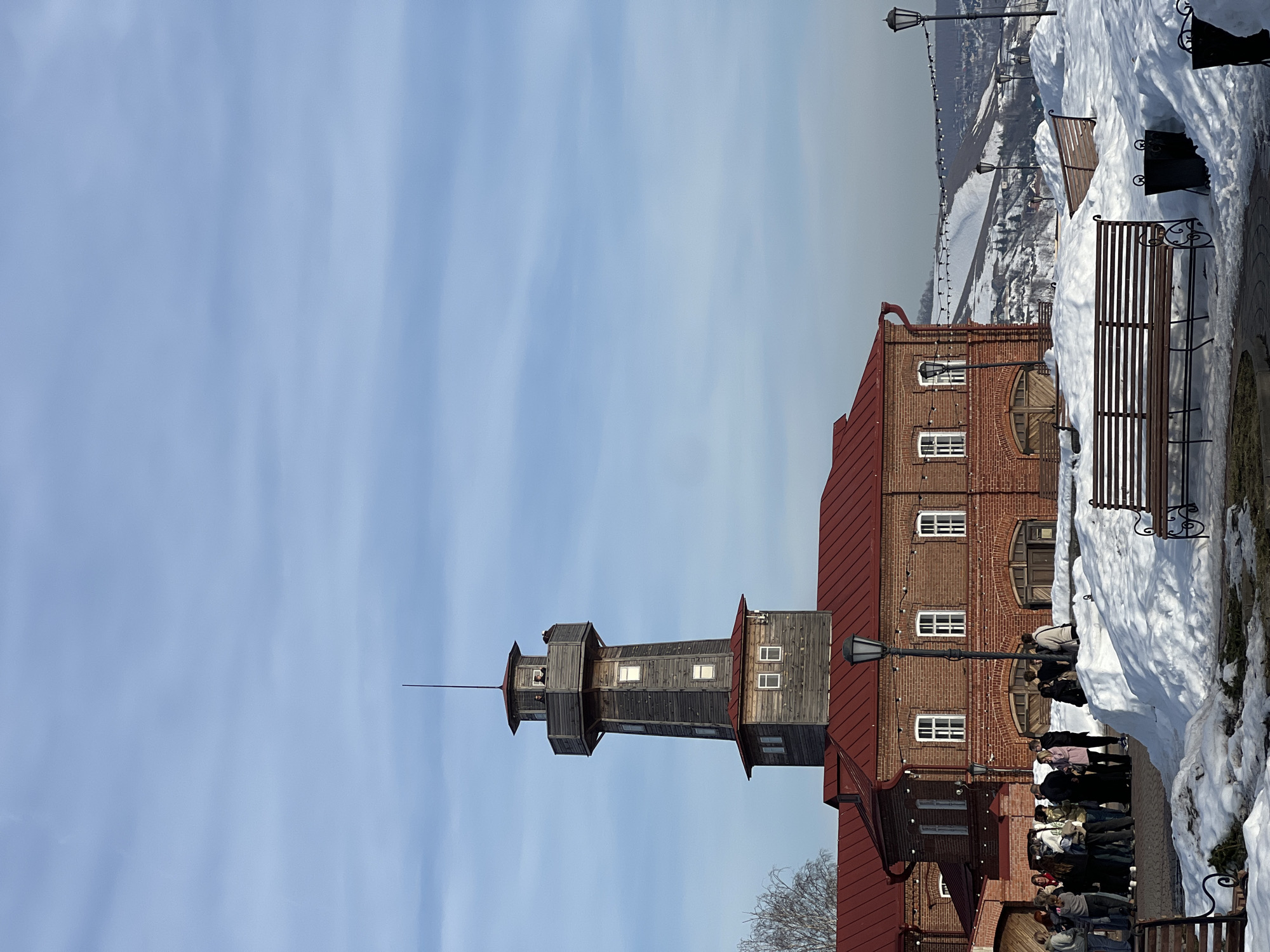 The image depicts a winter scene in Sviyazhsk, Russia, under a clear, pale blue sky with faint cirrus clouds. The central subject is a historic two-story red brick building with a red corrugated metal roof, featuring multiple white-framed rectangular windows and arched wooden door entrances. A prominent multi-tiered wooden fire lookout tower, constructed of weathered gray timber, rises from a section of the red roof. The tower has small square windows and an observation deck near its apex, where two individuals are visible. A slender metal spire extends from the tower's roof.
At ground level, the area surrounding the building is covered in patches of white snow, with clear pathways of cobblestone or paving visible. Approximately fifteen to twenty individuals, dressed in winter attire including dark jackets and hats, are congregated near an entrance on the left side of the brick building, appearing to be standing and conversing. Several empty wooden park benches with black metal frames are positioned across the snowy foreground and mid-ground, some partially obscured by snowdrifts. Black ornate streetlights and metal waste bins are also present in the snowy landscape.
In the background, snow-covered hills with sparse trees and scattered structures are visible under a hazy sky. A string of festive lights is discernible strung across the mid-ground landscape, leading towards the distant hills. The overall lighting suggests a bright, sunny winter day.