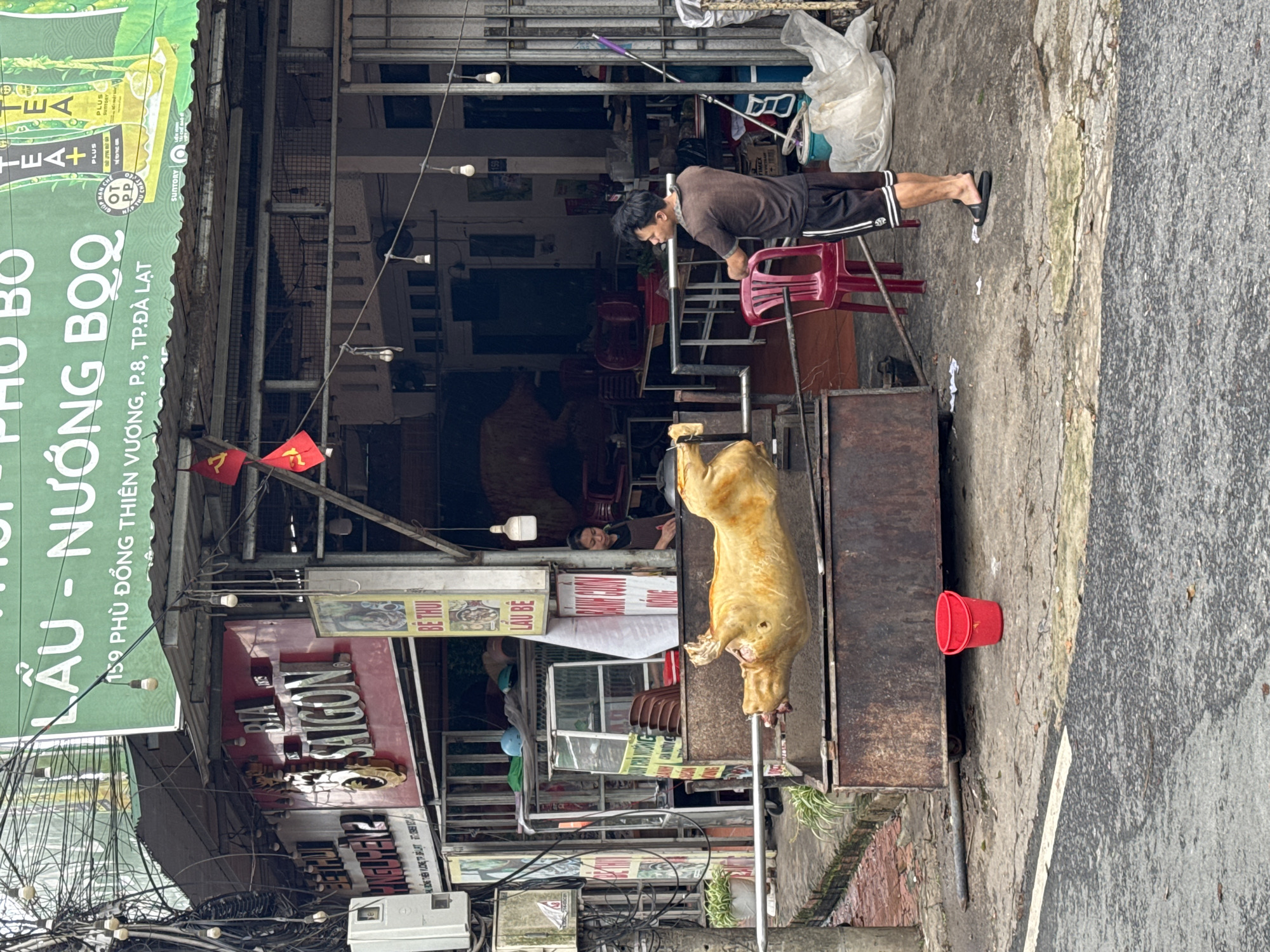 The image depicts a street scene in Đà Lạt, Lâm Viên province, Vietnam. A whole pig is being roasted on a spit in front of a restaurant. A man wearing a brown shirt and shorts is standing on a red plastic chair, appearing to tend to the roasting pig. The restaurant's sign reads "LẦU - NƯỚNG BQQ" and "BIA SAIGON," with other Vietnamese text visible. A person is sitting in the restaurant, behind the pig. Electrical wires are tangled above the street. A red plastic bucket is next to the roasting spit.