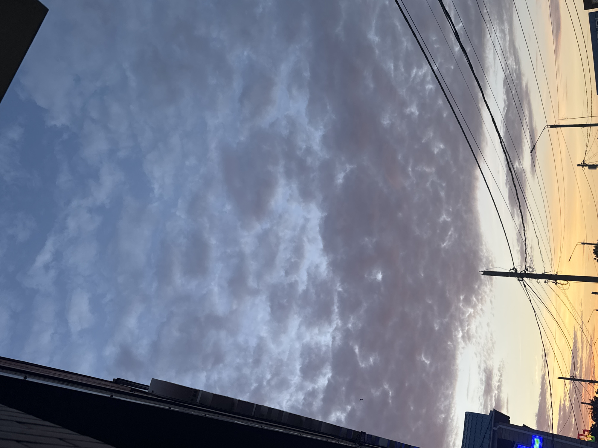 This is a photograph of a sky with dramatic clouds, likely taken at sunset or sunrise. The sky transitions from a pale blue with scattered, textured clouds on the left to a warm, golden hue with wispy clouds on the right. A few dark, silhouetted buildings and rooftops are visible at the bottom of the frame, suggesting an urban or semi-urban setting. Power lines and utility poles stretch across the upper portion of the image, indicating the presence of infrastructure. The overall mood is serene and atmospheric, with the warm tones of the sky creating a sense of peace. The image appears to be taken from a low angle, looking upwards towards the sky. There is no visible text in the image.
