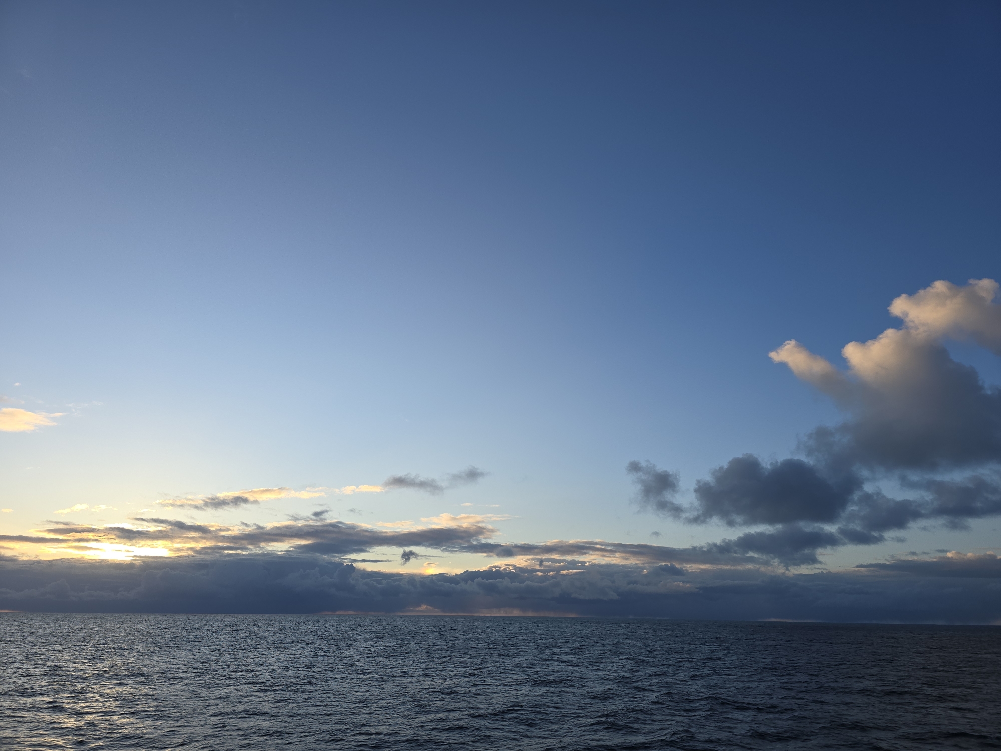 The image shows a body of water, likely an ocean, under a sky with varying cloud cover. The clouds near the horizon are thicker and darker, obscuring a portion of the setting sun. Above these, the sky transitions to a clear, deep blue. To the right, a few puffy white clouds stand out against the blue. The water's surface is rippled, suggesting a slight breeze. The location in the city is unknown.