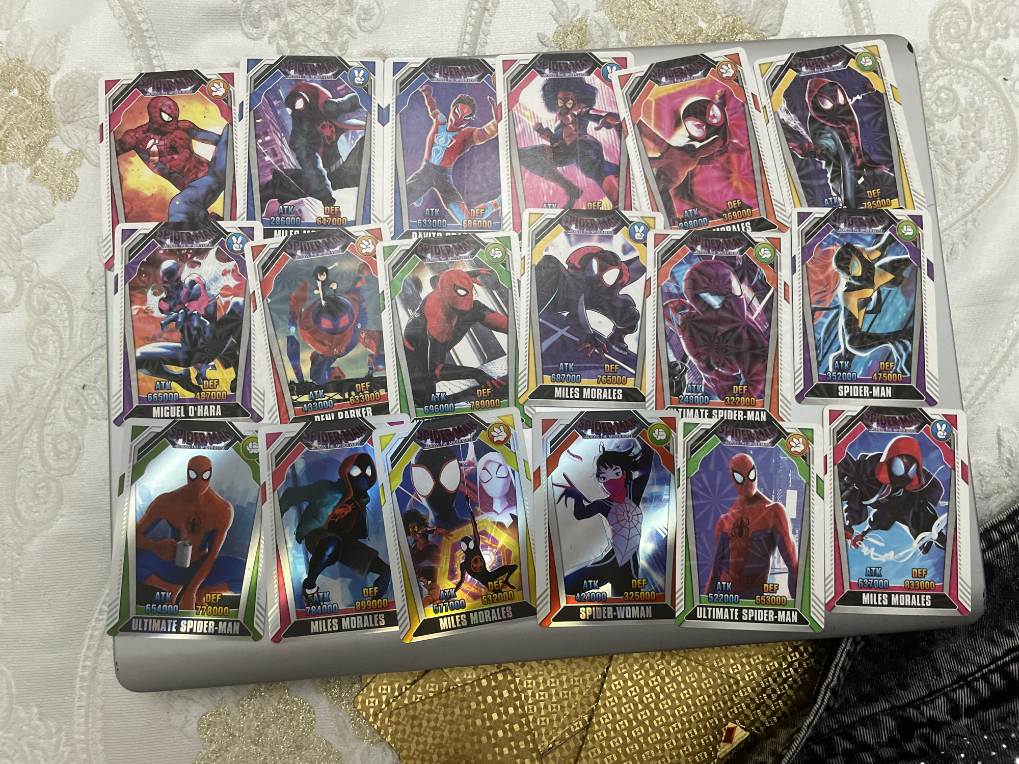 A collection of approximately eighteen Spider-Man trading cards from "ACROSS THE SPIDER-VERSE" is spread across a light-colored tablecloth, possibly resting on a closed silver or gray laptop. The tablecloth features an intricate, light gold patterned design. Each card showcases a different Spider-person or variant, rendered in dynamic, comic-book-style art. Characters visible include multiple versions of Miles Morales, Miguel O'Hara (Spider-Man 2099), Ultimate Spider-Man (Peter Parker, including one holding a mug), and Spider-Woman.
Every card displays the character's name, "ATK" (Attack) and "DEF" (Defense) statistics with various numerical values (e.g., ATK 654000 DEF 778000 for Ultimate Spider-Man; ATK 286000 DEF 647000 for Miles Morales). Several cards also feature a small symbol in the top right corner, such as a rabbit head, a paw print, or a leaf icon. The cards are brightly colored and glossy, with some overlapping slightly. The arrangement suggests they are being displayed or sorted.