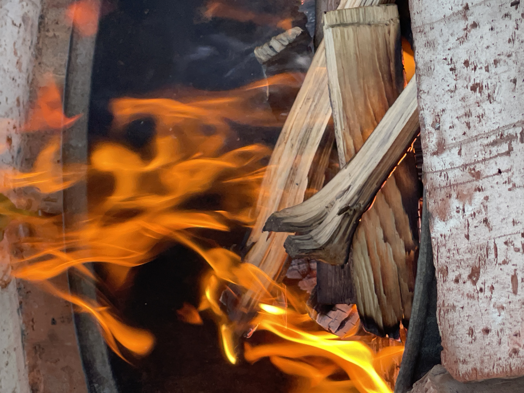 The image shows a close-up view of a fire burning in what appears to be a fire pit or outdoor oven. Several pieces of firewood are stacked inside, with bright orange flames engulfing some of the wood. The fire is intense, with dynamic flames reaching upwards. The edges of the fire pit are visible, made of brick or stone, with some signs of wear and tear. The background is dark, suggesting the image was taken in low light conditions or the fire is contained within a darker space. No people are visible in the image. The scene depicts a controlled fire for warmth or cooking, with the primary focus on the active flames and burning wood. The location in Orenburg, Russia, is not discernible from the visual information provided in the image.