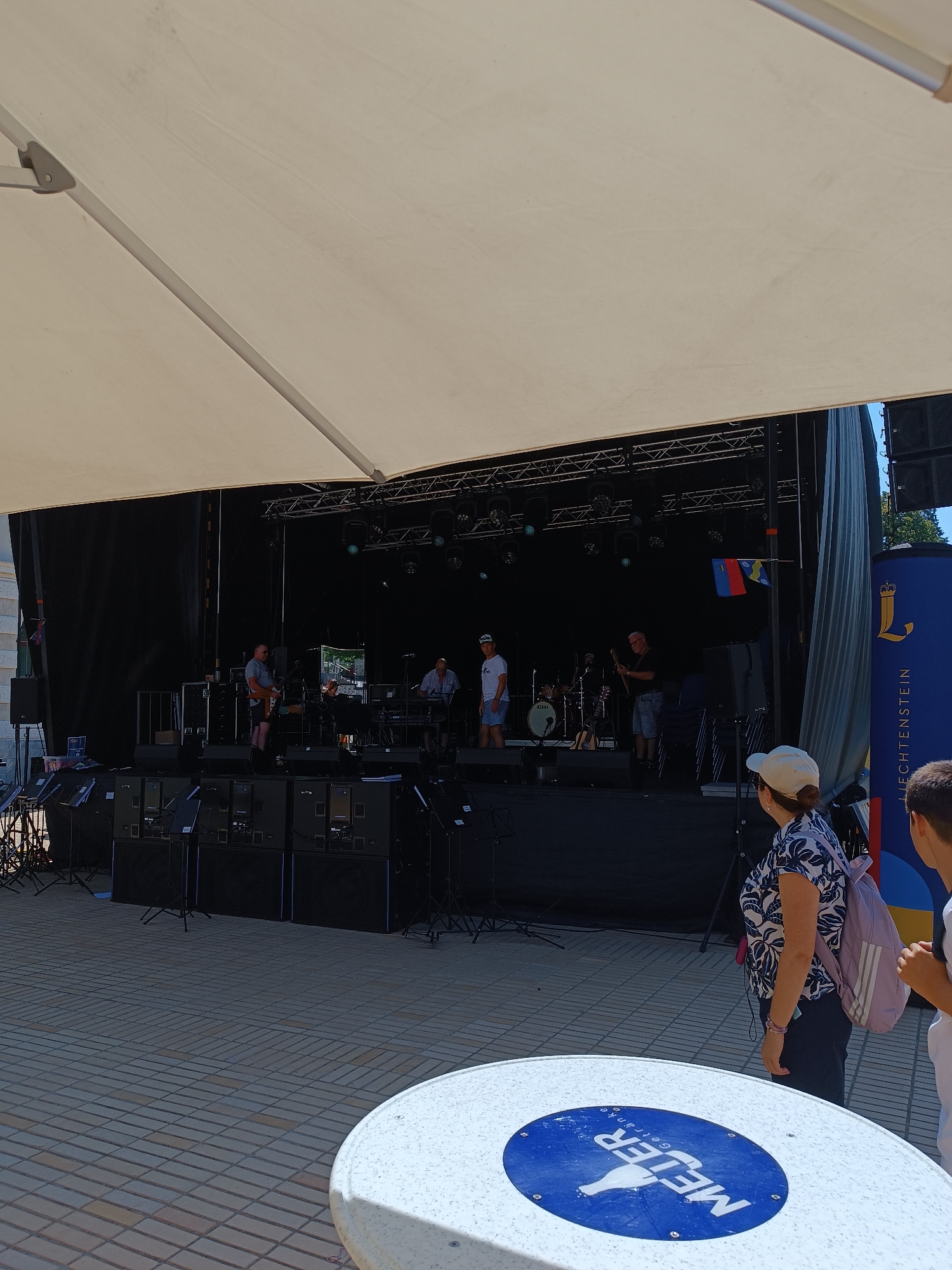 The image captures an outdoor event with a band performing on a stage. A white canopy covers the stage area, casting a shadow over the foreground. The stage is set up with musical equipment, including speakers, amplifiers, and instruments. Several band members are visible on stage, playing guitars and drums. In the background, a large banner with the words "LIECHTENSTEIN" and a royal emblem suggests the location is Vaduz, the capital of Liechtenstein. A small flag with the colors red, blue, and yellow is also displayed. In the foreground, a woman wearing a patterned shirt and a backpack stands facing the stage, along with another person partially visible. A white circular table with a blue logo is in the immediate foreground, indicating a public seating area. The ground is paved with interlocking square bricks.