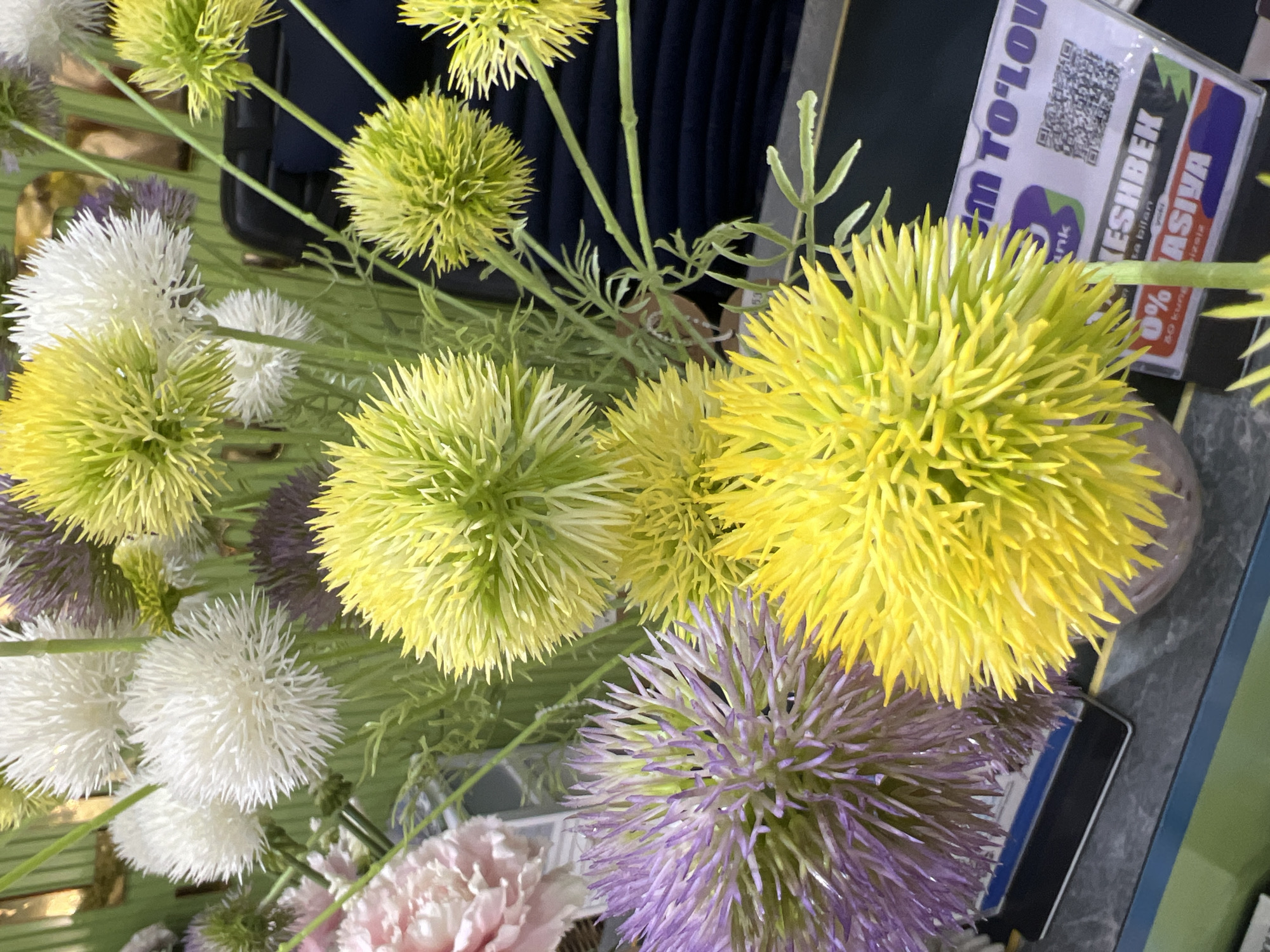 The image shows a close-up view of an artificial floral arrangement featuring spiky, globe-shaped flowers in shades of yellow, purple, and white, along with some pale pink petals from a different flower. The flowers are densely packed and appear to be made of plastic or a similar material, with thin, pointed petals radiating outwards from a central point. Green, feathery foliage is interspersed throughout the arrangement. In the background, dark blue fabric and what appears to be a hint of a metallic gold object are visible. A sign with text and a QR code is positioned in the lower right portion of the frame. The sign appears to have some text in a foreign language and mentions "EM TO'LOU" and "ASHBEK NASIYA". This detail and the specific mention of Eshanguzar, Uzbekistan in the prompt suggest the image was taken in a commercial setting within that city, possibly a shop or market stall selling artificial flowers or related items. There are no people visible in the image.