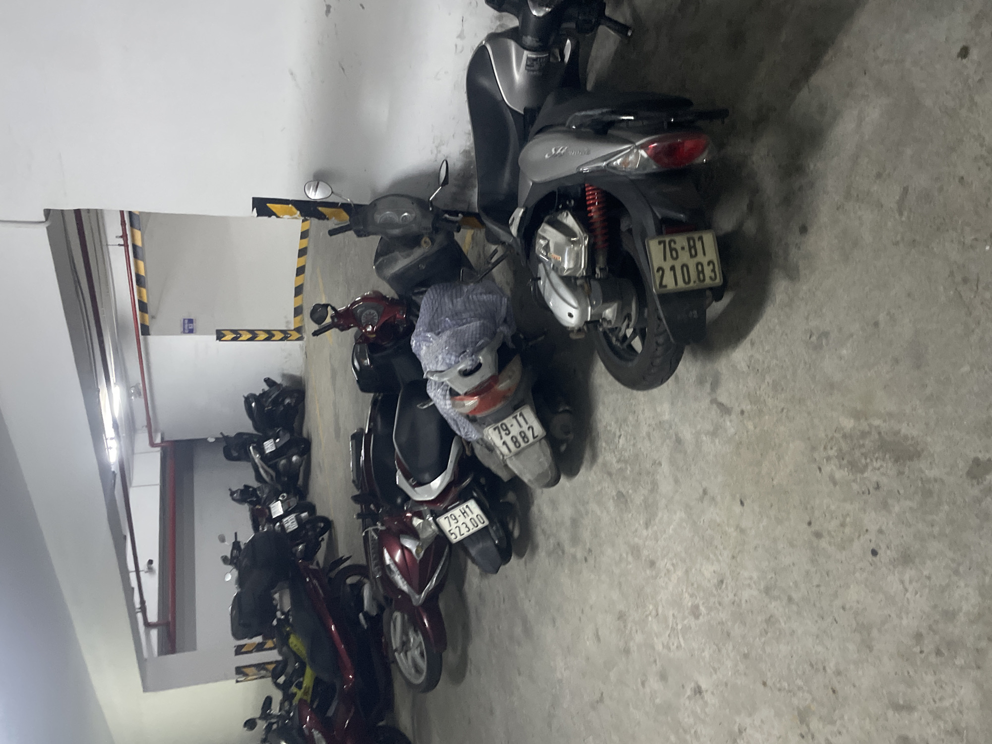 The image shows a parking garage in Bac Nha Trang Ward, Vietnam, filled with parked motorcycles. In the foreground, a silver-grey scooter lies on its side. To its left, a maroon scooter is also on its side, with a white and blue cloth draped over its seat. Further to the left, another maroon scooter is parked upright, and behind it, several other motorcycles of various colors are visible. The garage has a concrete floor and white walls with red and yellow striped markings indicating caution. Lights are visible on the ceiling. No people are present in the image.