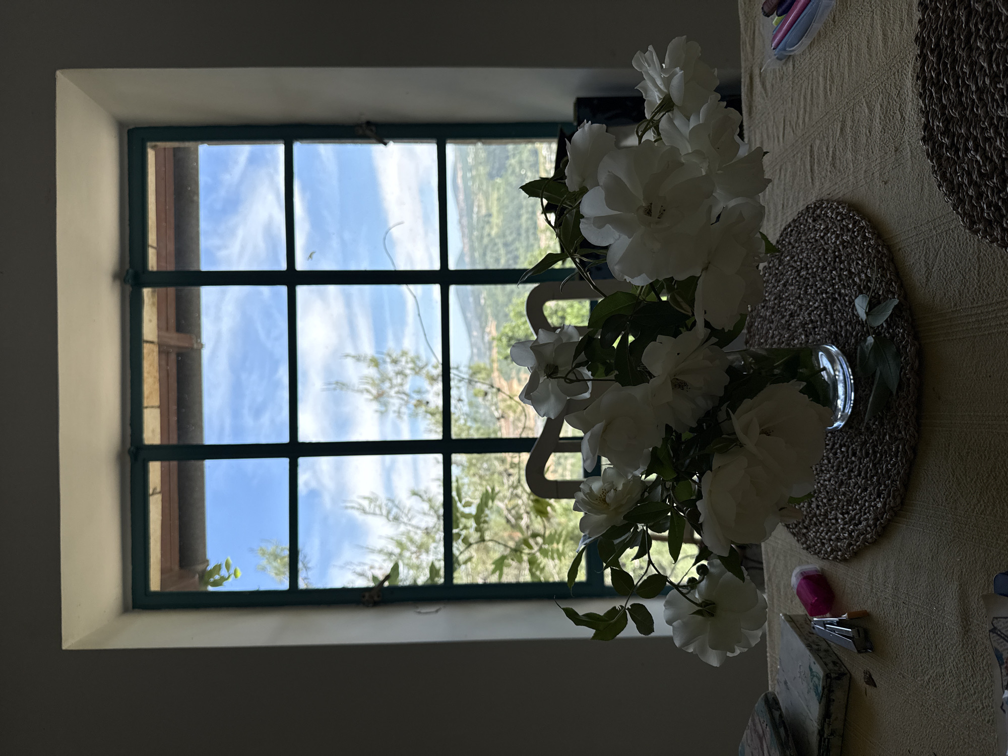 The photograph depicts an indoor scene featuring a window and a table setting. The window, divided into multiple panes with a dark green frame, looks out onto a landscape with vegetation, a clear blue sky, and distant hills that indicate the scene is in Kfar Vradim, Israel. A vase of white flowers is placed to the right of the window, partially obscuring the view. The table is covered with a textured beige cloth and has a braided placemat. Other objects, including a pink eraser, stapler, and various items, are on the table. The room itself appears dimly lit, and the focus is on the contrast between the indoor space and the bright outdoor scenery.