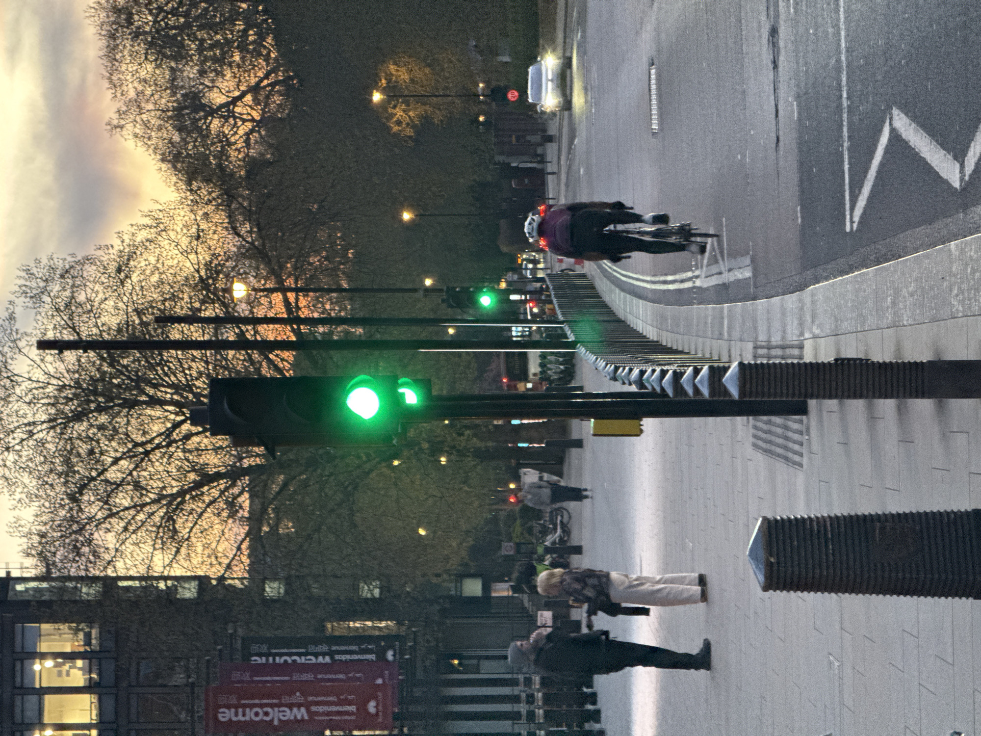 The photograph, taken in the city of London, United Kingdom, shows a street scene at dusk. A cyclist wearing a helmet and dark clothing is riding on the road, passing a traffic light that is showing a green light. A white vehicle is also visible further down the road. On the pavement, pedestrians are walking, including a person wearing light-colored trousers and a person wearing a dark coat. There are black metal barriers separating the road from the pavement. A building with "Welcome" signs is partially visible on the left side of the frame. The sky behind the trees shows sunset colors.