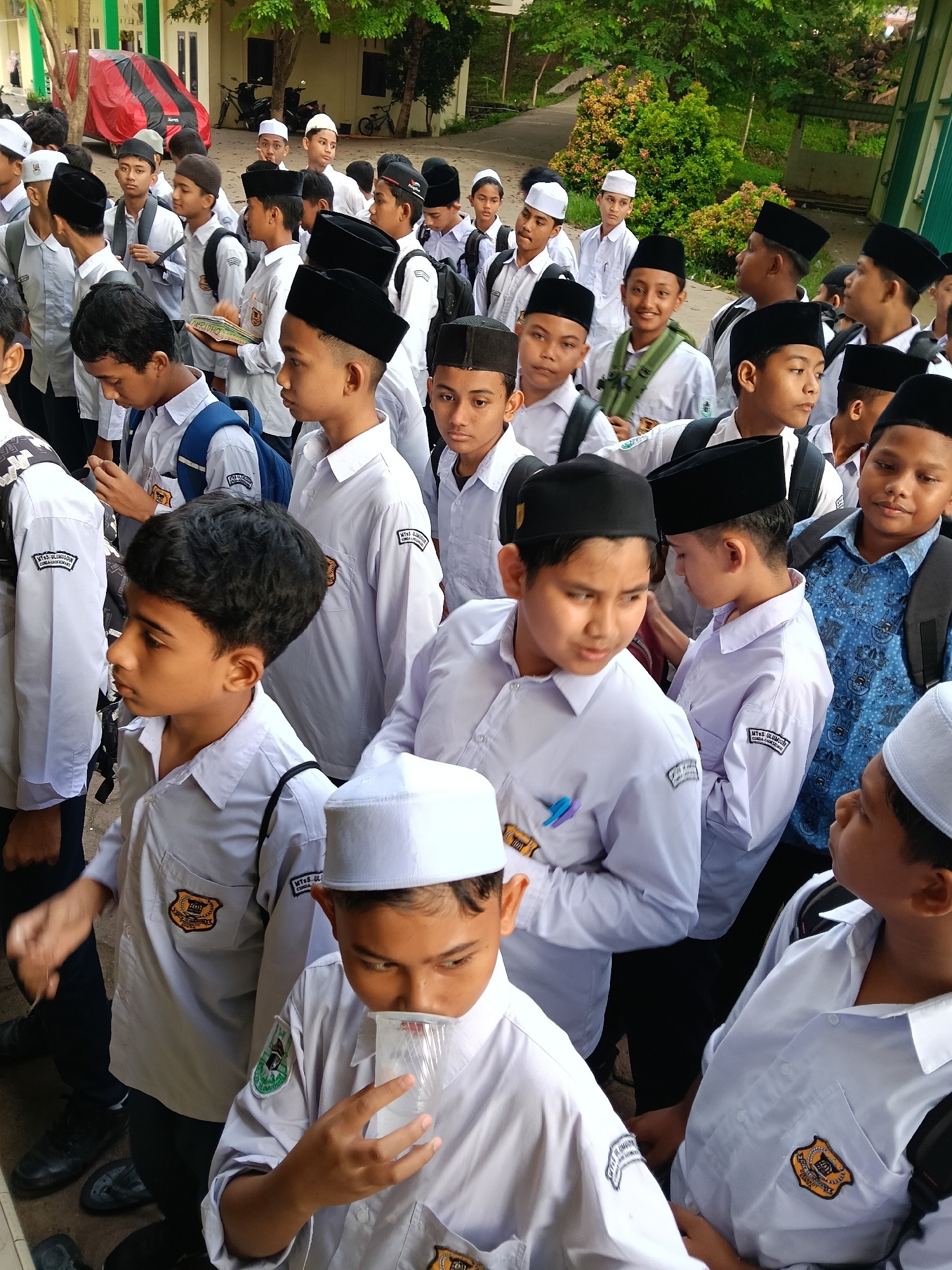 A medium, eye-level shot depicts a dense group of young male students, predominantly wearing white collared shirts with distinct shoulder patches and emblems on their chests. Many are sporting black peci hats, though some have white ones. Several have backpacks. The scene appears to be set outdoors, possibly on school grounds. A boy in the foreground is drinking from a clear plastic cup. This photograph was taken in the city of Lhokseumawe, Indonesia.
