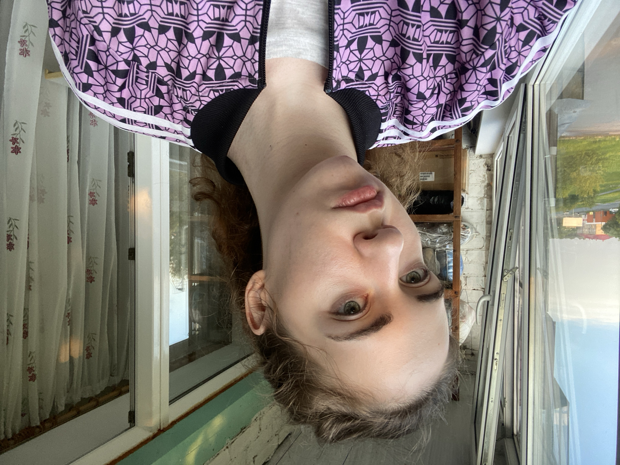 The media file is an upside-down, close-up, portrait-style photograph of a young person with light skin and green eyes. They are wearing a purple jacket with a geometric black and white pattern, and a grey t-shirt underneath. The background includes a window with white curtains that have a floral pattern, a brick wall, and a view of a field and some buildings outside. The lighting suggests it is daytime, possibly late afternoon or early evening, with a soft, natural light. The overall mood appears casual and relaxed, with no strong emotions conveyed. The person's face is the primary focus, and their gaze is directed towards the camera. There is no visible text in the image. The context provided indicates the location is Zavodoukovsk, Russia.