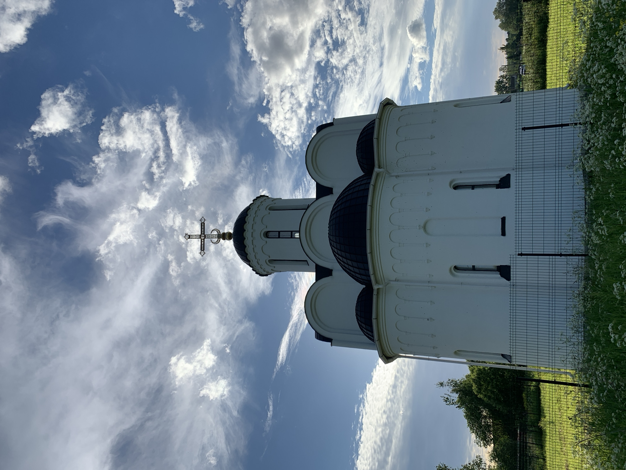 The photograph captures a white, traditional Russian church with a black, domed roof, set against a backdrop of a blue sky with wispy white clouds. A cross adorns the central dome. The church is surrounded by a green grassy area with scattered white wildflowers and a black metal fence in the foreground. Trees are visible in the background. The location is specified as Новый Учхоз, Russia. There are no people visible in the image.