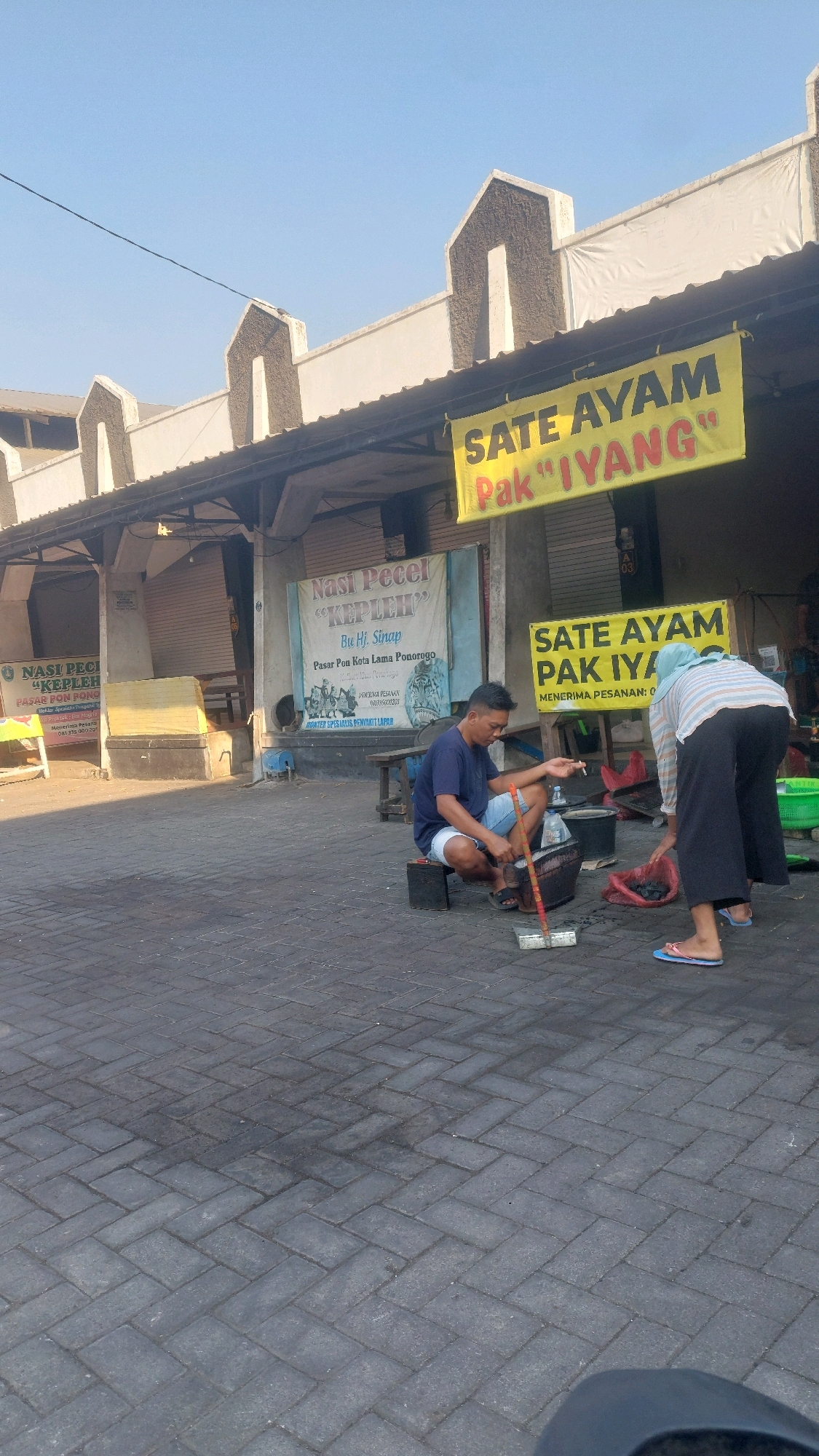 The image captures a street scene in Nologaten, Indonesia, with a focus on a food stall area. In the foreground, a man wearing a dark blue t-shirt and light blue shorts is squatting down, appearing to be preparing charcoal for a grill. He holds a long stick and is positioned next to a black bucket. To his right, a woman wearing a light-colored striped shirt and dark pants, a headscarf, and flip-flops is also visible, tending to a red bag containing charcoal. Behind them, several food stalls are set up, indicated by signs. One prominent yellow banner reads "SATE AYAM PAK 'IYANG'". Another banner advertises "Nasi Pecel 'KEPLEH'". The ground is paved with dark grey interlocking bricks. The overall environment suggests an outdoor market or a row of shops on a sunny day. The architecture of the buildings features a distinctive angular roofline.