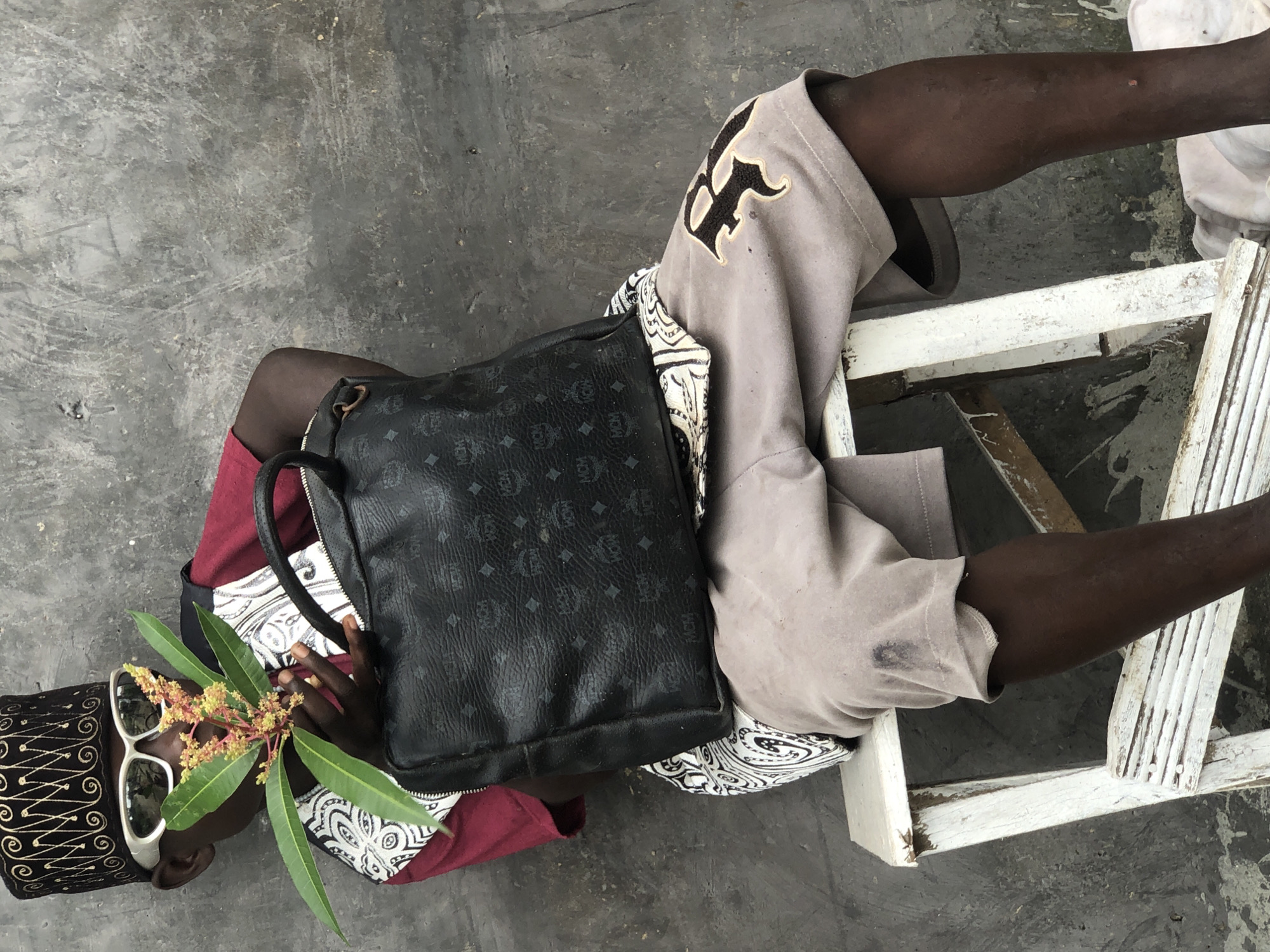 A dark-skinned person wearing a black and gold embroidered hat, white sunglasses, a red shirt, a patterned vest, and beige shorts with a black "R" emblem sits on a white wooden stool against a grey concrete wall. The person holds a black designer handbag with a patterned logo. In their hands, they hold a small flowering branch with green leaves and yellowish-orange flowers. The scene appears to be outdoors in a city, possibly Ado, Nigeria.