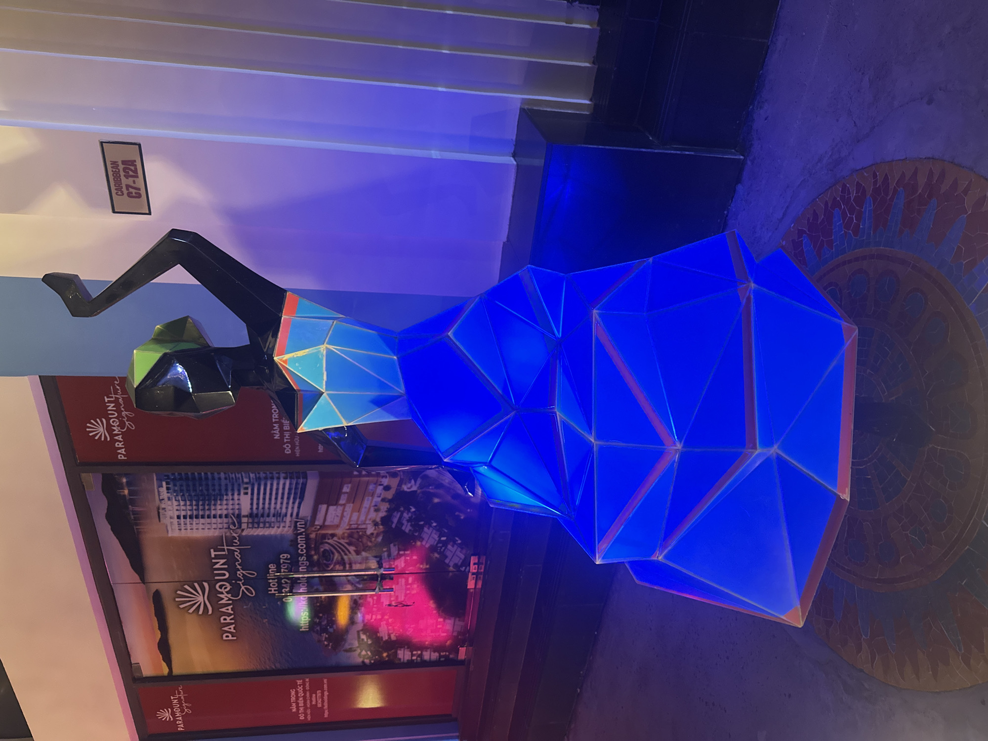Here is a description of the image:
The photograph shows a modern art sculpture of a stylized human figure in Bac Nha Trang Ward, Vietnam. The sculpture appears to be made of geometric shapes and is lit up with a bright blue light. It is placed in front of a promotional display for "Paramount Signature," a resort or residential development. The display features images of buildings and a coastline at sunset. The artwork is partially obscured by the "Paramount Signature" marketing panels. A wall and floor with mosaic design are also visible in the background. A sign on the wall reads "CARIBBEAN C7-12A".