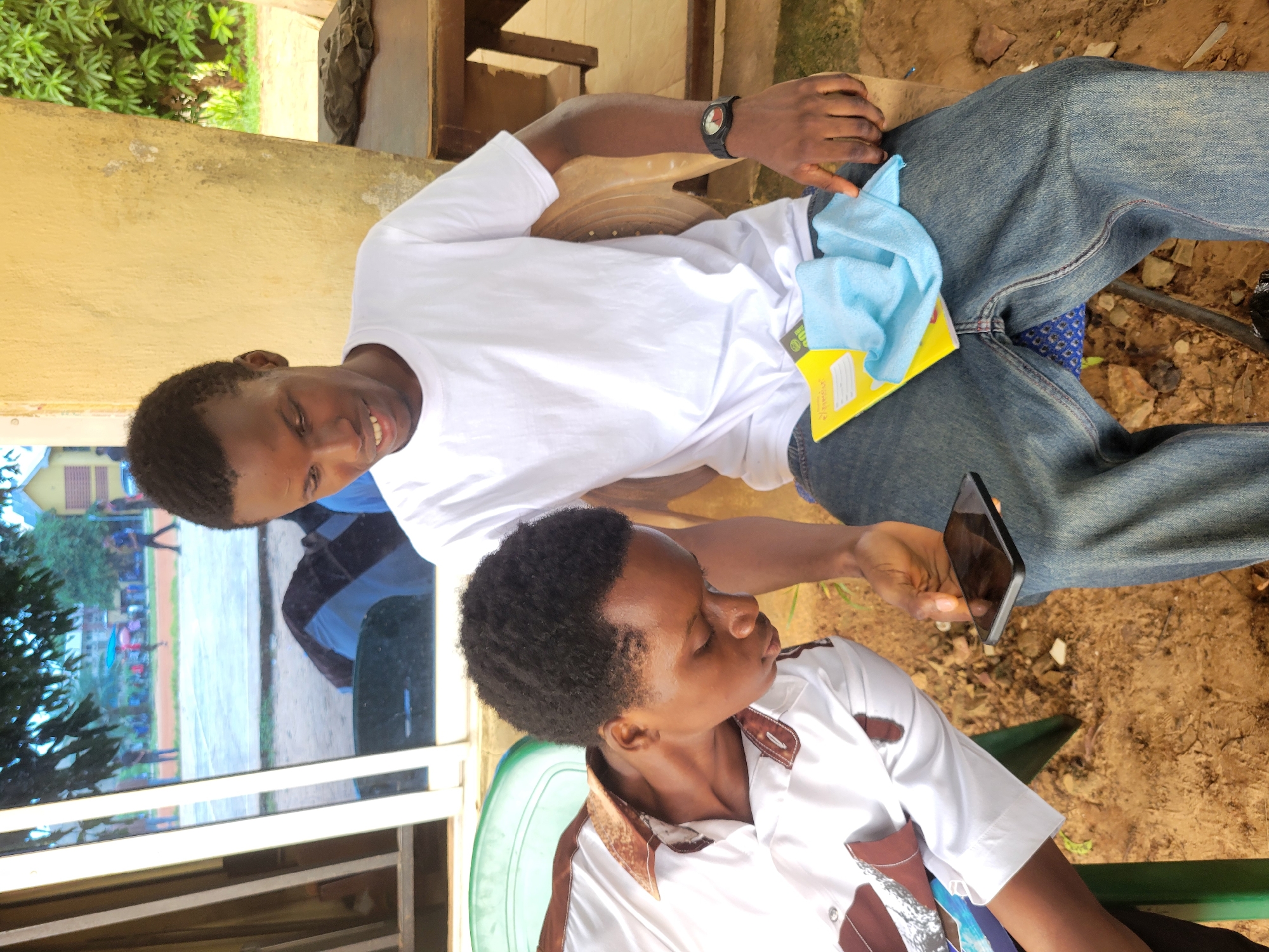 Two young men are seated outdoors. The man on the left is wearing a white shirt with brown trim and is looking down at a smartphone he holds in his right hand. The man on the right is wearing a white t-shirt and blue jeans and is looking at the phone as well, with his right hand resting on his thigh. A yellow notepad and a light blue cloth are on his lap. The background shows a building with yellow walls and green foliage, and in the distance, a basketball court with people in it. The scene appears to be in the city of Ukukwa, Nigeria.