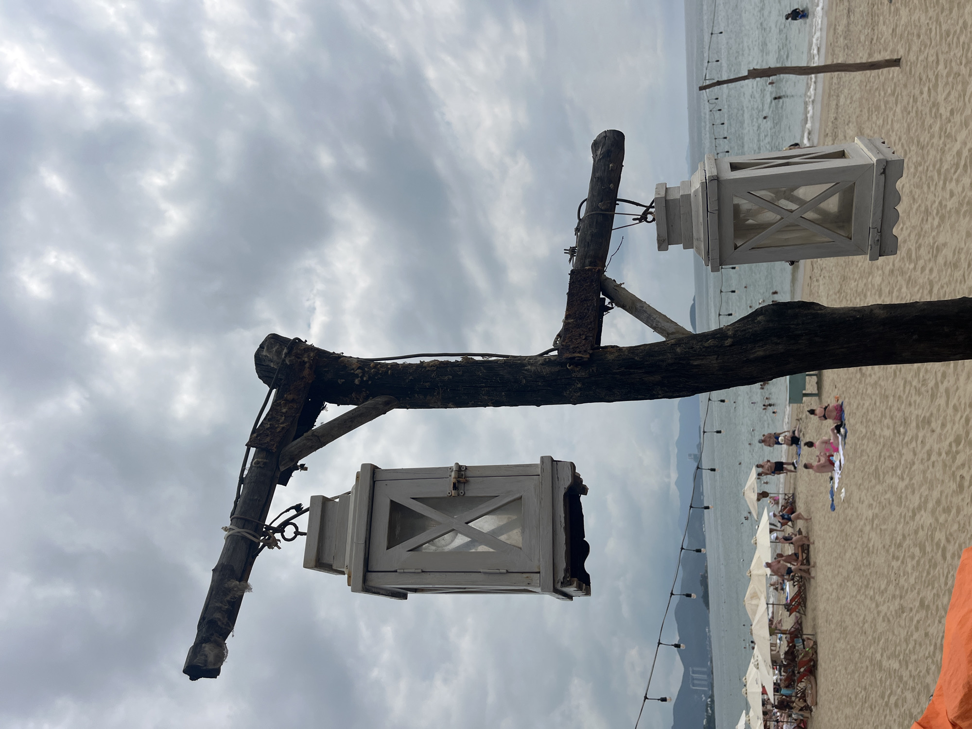 The photograph shows a beach scene in Nha Trang, Vietnam, featuring two white, wooden lanterns hanging from a weathered wooden structure. The structure consists of a vertical post with a horizontal branch and a support brace. Each lantern has an X-shaped design on its sides and is suspended by metal hooks. In the background, the beach is populated with people swimming, sunbathing, and relaxing under white umbrellas. Mountains are visible in the distance across the water. A string of lights hangs along the edge of the frame. The sky is overcast with grey clouds.