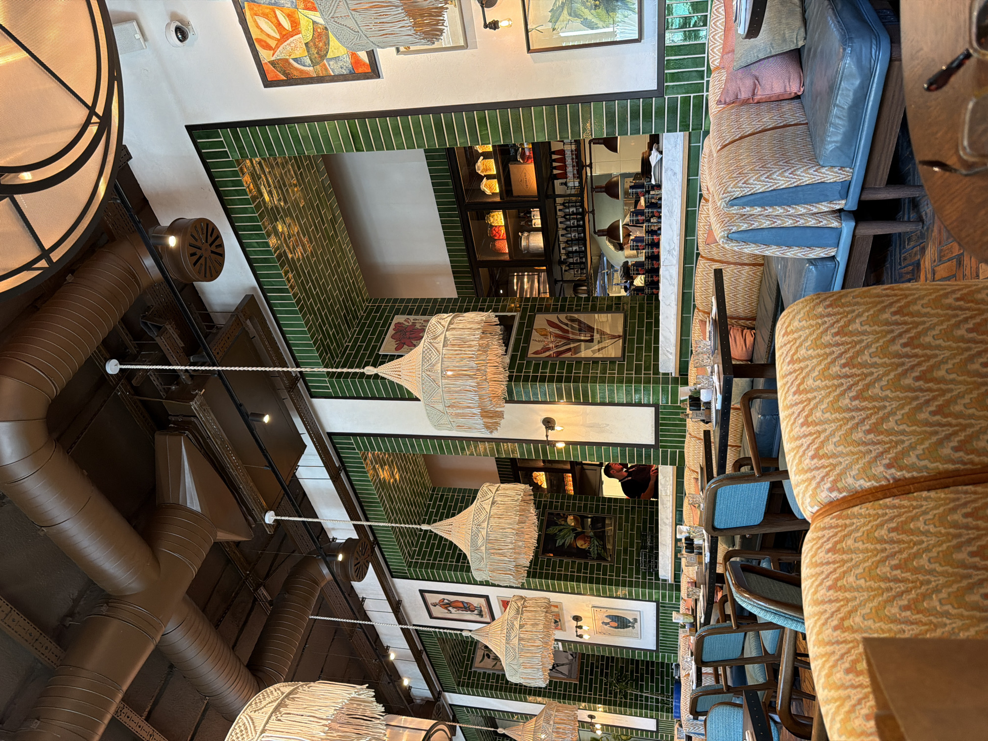 This image depicts the interior of a restaurant with a vibrant and eclectic design. The dominant color scheme includes shades of green, orange, and blue. The restaurant features numerous decorative elements, including macrame lampshades hanging from the ceiling, framed artwork on the walls, and plush seating upholstered in patterned fabrics.
The walls are covered in green subway tiles, creating a striking visual contrast with the exposed brown ductwork visible on the ceiling. The seating arrangements include booths with blue leather upholstery and colorful patterned cushions, as well as individual chairs. Tables are set with tableware, suggesting an active dining environment.
In the background, a bar area is visible, with shelves stocked with bottles and jars. A person in a dark uniform can be seen behind the counter, indicating staff presence. The lighting is a mix of ambient overhead lights and spotlights, contributing to the warm and inviting atmosphere.
While the image captures the interior of a restaurant, it does not contain specific visual cues that would allow for its precise location within Sabail Raion, Azerbaijan. The general aesthetic is consistent with modern restaurant design and could be found in various urban settings.