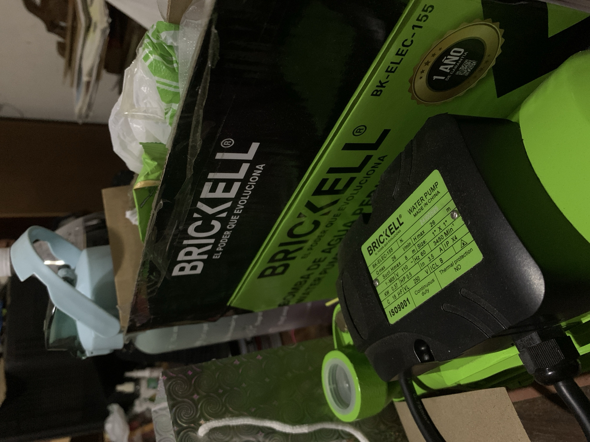 The image captures a close-up, angled view of a green and black "BRICKELL" water pump, partially nestled within its packaging and surrounded by other household items in what appears to be an indoor storage setting. No people are visible, and no specific activity is taking place; the scene suggests items are either stored or recently acquired.
The "BRICKELL" brand name is prominently displayed on both the pump's label and its cardboard packaging, along with the slogan "EL PODER QUE EVOLUCIONA" (The Power That Evolves). The product is identified as a "WATER PUMP" ("BOMBA DE AGUA") with model number "BK-ELEC-155." A gold warranty sticker on the packaging states "1 AÑO DE GARANTÍA" (1 YEAR WARRANTY) and features a QR code.
The green product label on the pump provides detailed specifications, including "MADE IN CHINA," power rating "KW 0.37 HP 0.5," voltage "110~Hz 60," and an "IP X4" rating. It also mentions "IS09001" certification and "Continuous duty" with "Thermal protection NO." Other numerical specifications like "Qmax 26," "H max 8 m," and "Suct.Hmax 26 m" are also listed.
Other visible items include a crumpled white plastic bag, a light blue plastic object that resembles a handle or part of a small appliance, and hints of cardboard boxes and other clutter in the dimly lit background. The text in Spanish and the provided location context of Santa Ana, Venezuela, suggest the market where the product is sold. The time of day is indeterminate due to the indoor setting.